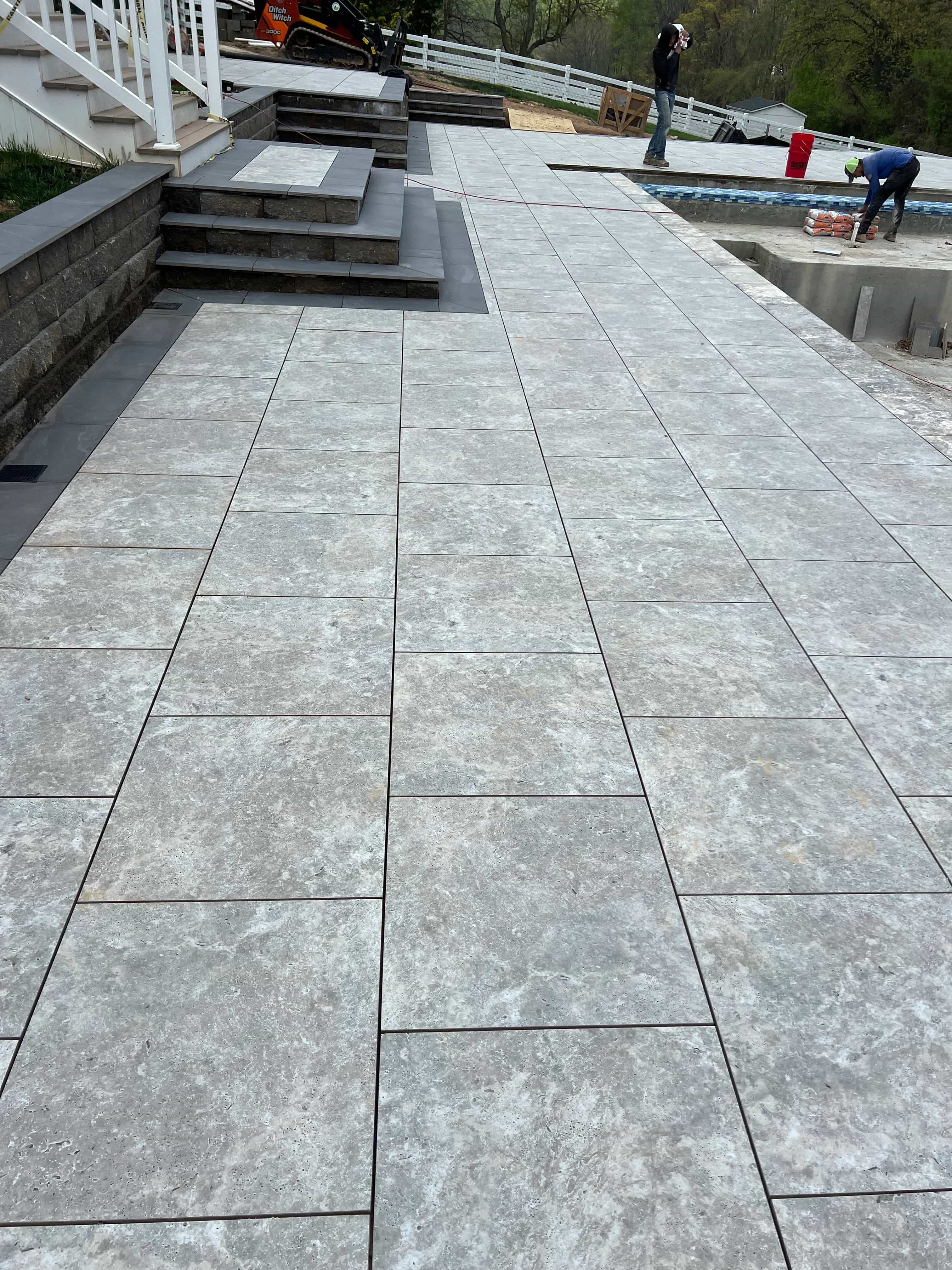 Installation of Travertine Silver Matte 24x36x2cm Rectified Porcelain Paver by STONELUX, used for a modern patio. Features matte finish and natural stone look, complementing dark stone steps. Surrounded by white fencing and greenery, ideal for hardscap...