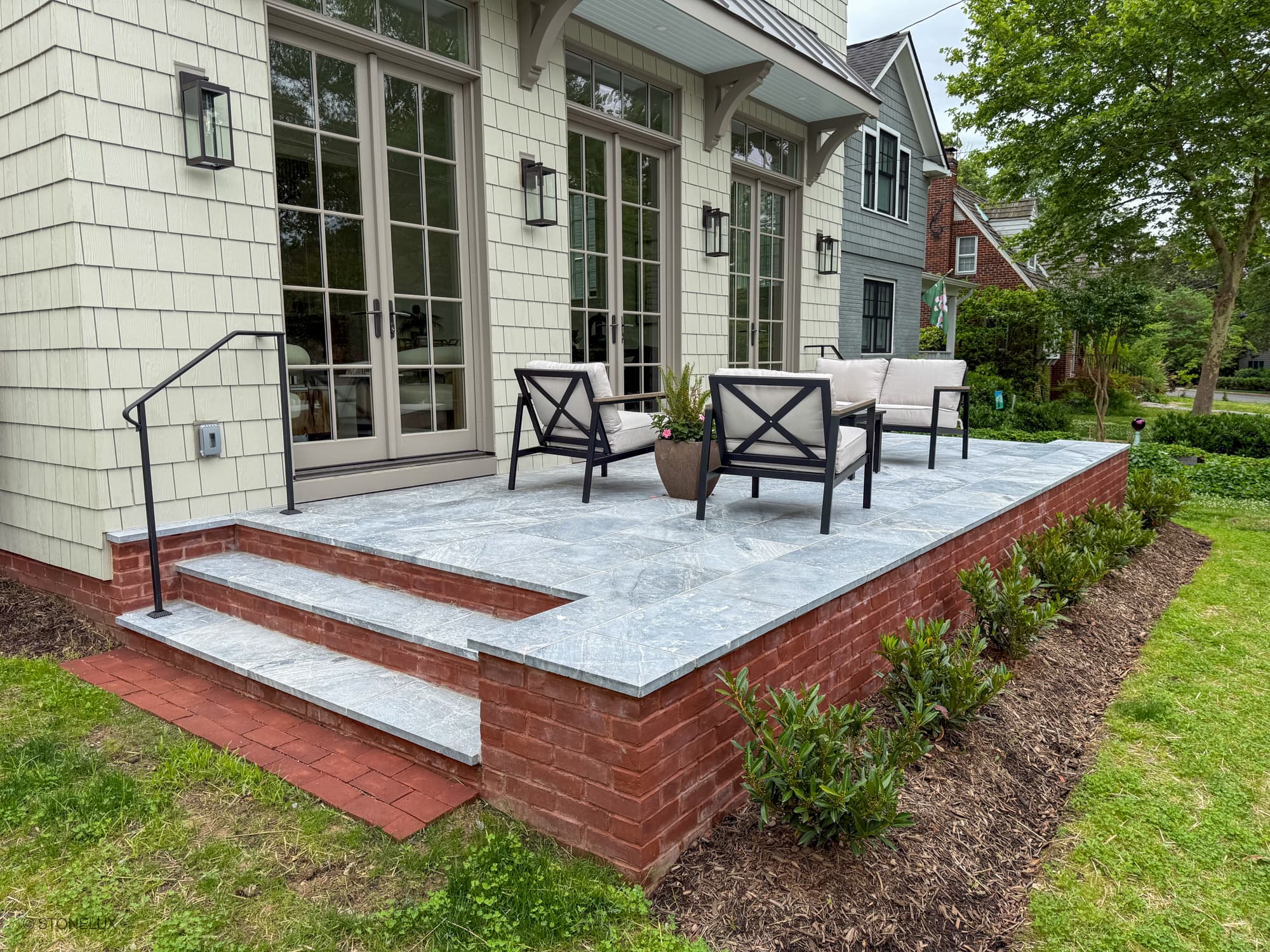Outdoor patio featuring Aegean Grey Marble Sand Blasted 16x24 3cm Paver by STONELUX. The elegant marble pavers are installed on a raised brick platform, complementing the traditional home architecture with modern outdoor seating and lush greenery.