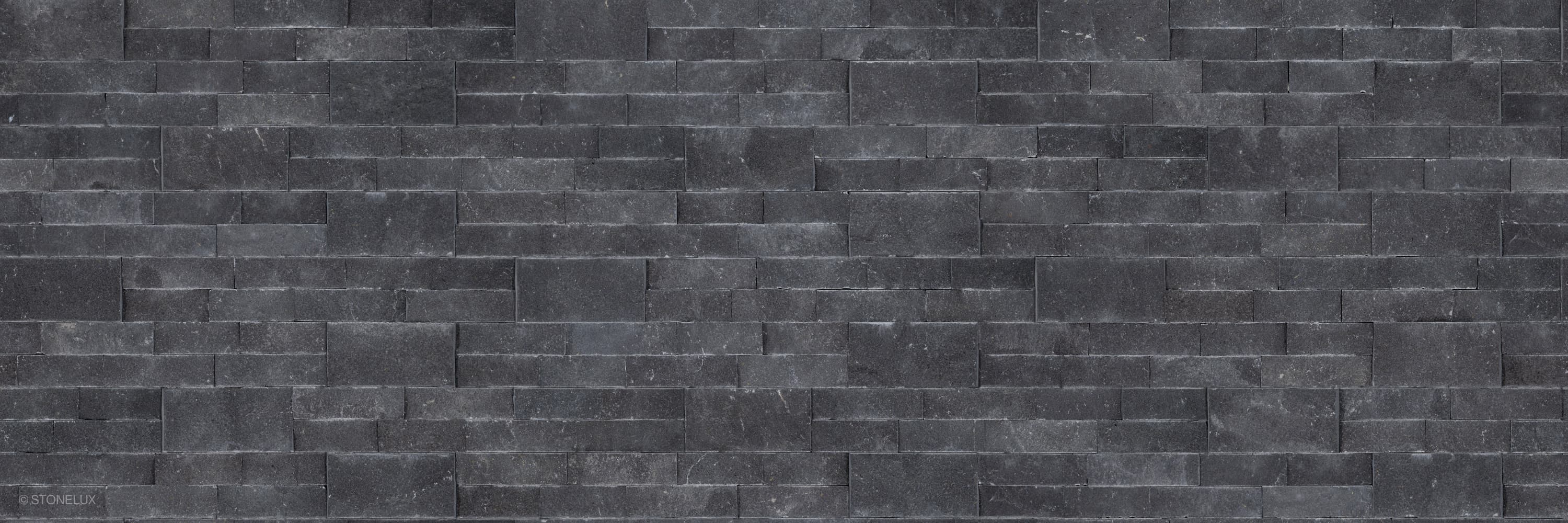 Close-up image of Black Basalt Splitface Ledger Panel - 6x24, showcasing its rugged splitface texture and deep charcoal hues. The natural basalt stone veneer features subtle variations and striations, ideal for modern wall installations and hardscape p...