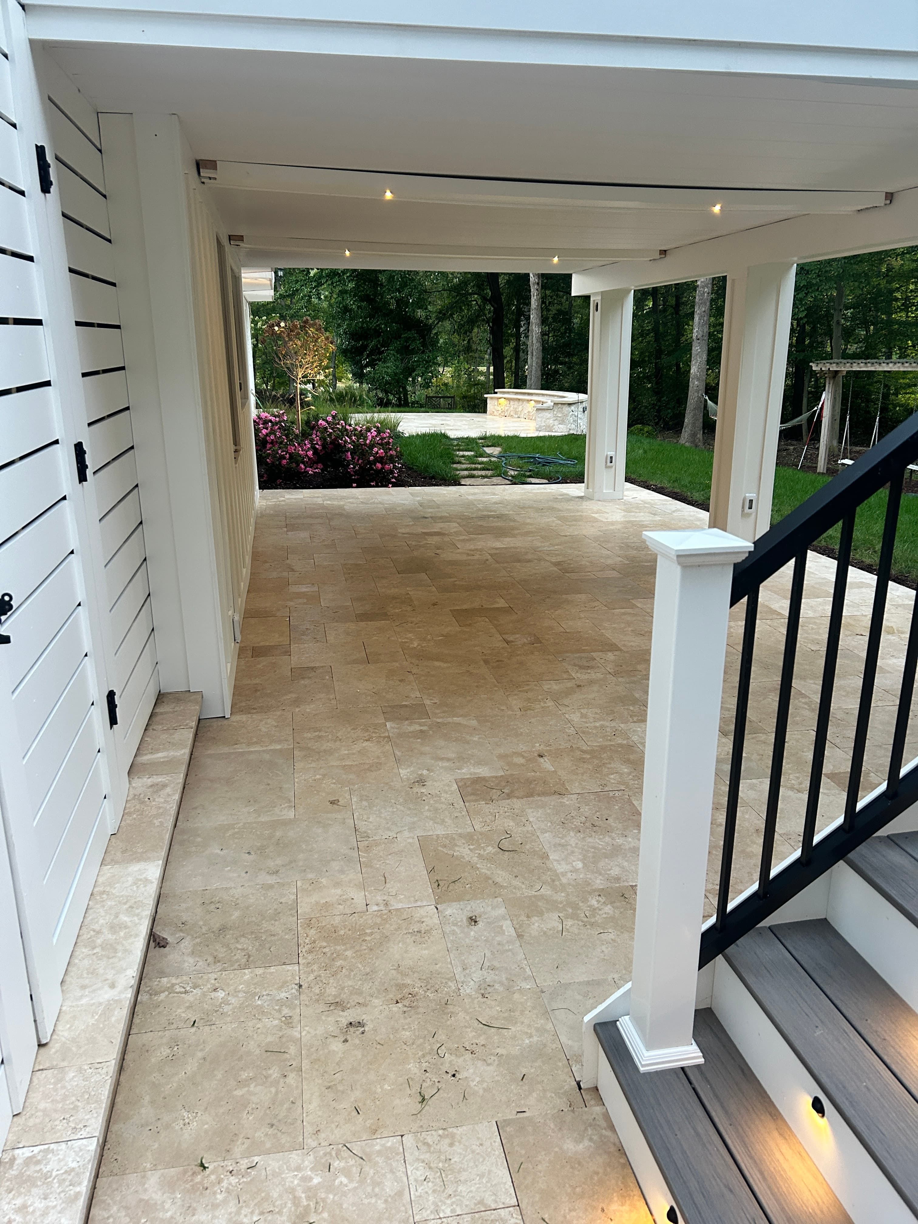 Covered patio featuring Ivory Travertine Tumbled French Pattern 3cm Paver - Premium by STONELUX. The natural stone flooring complements the modern white architecture and lush garden. Ideal for hardscape projects seeking elegance and durability.