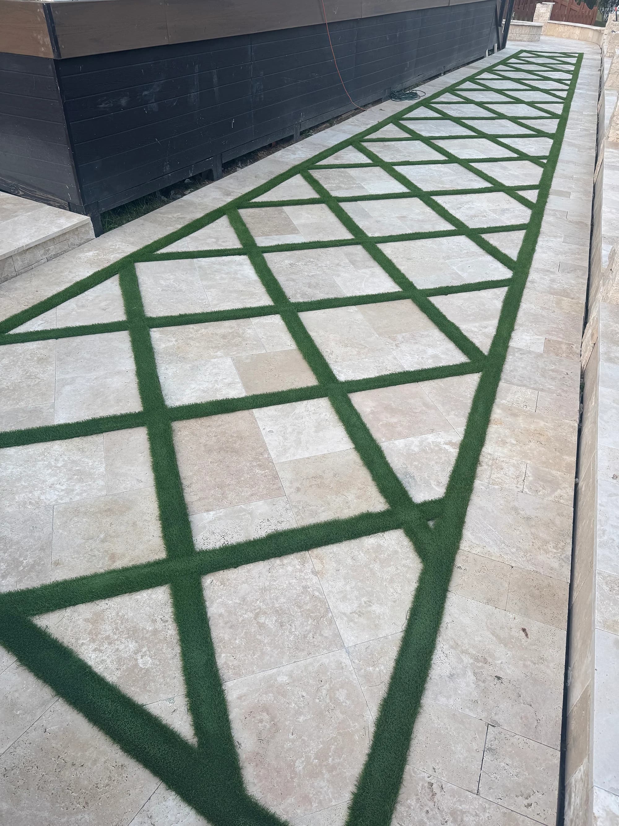 Ivory Travertine Tumbled French Pattern 3cm Paver installation by STONELUX. The natural stone features warm beige tones and a tumbled finish, beautifully arranged with green grass strips creating a striking pattern. Ideal for modern hardscape projects.