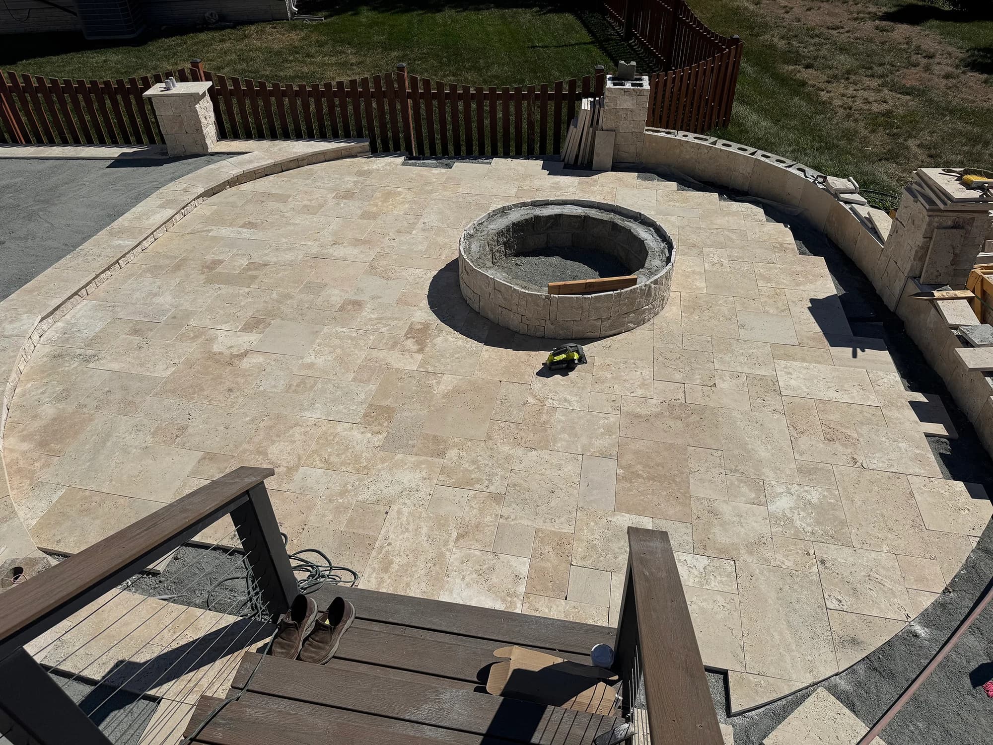 Ivory Travertine Tumbled French Pattern 3cm Paver creates a stunning patio installation. The natural stone's creamy tones and tumbled finish complement the curved design and central fire pit, bordered by rustic wooden fencing and lush greenery, ideal f...