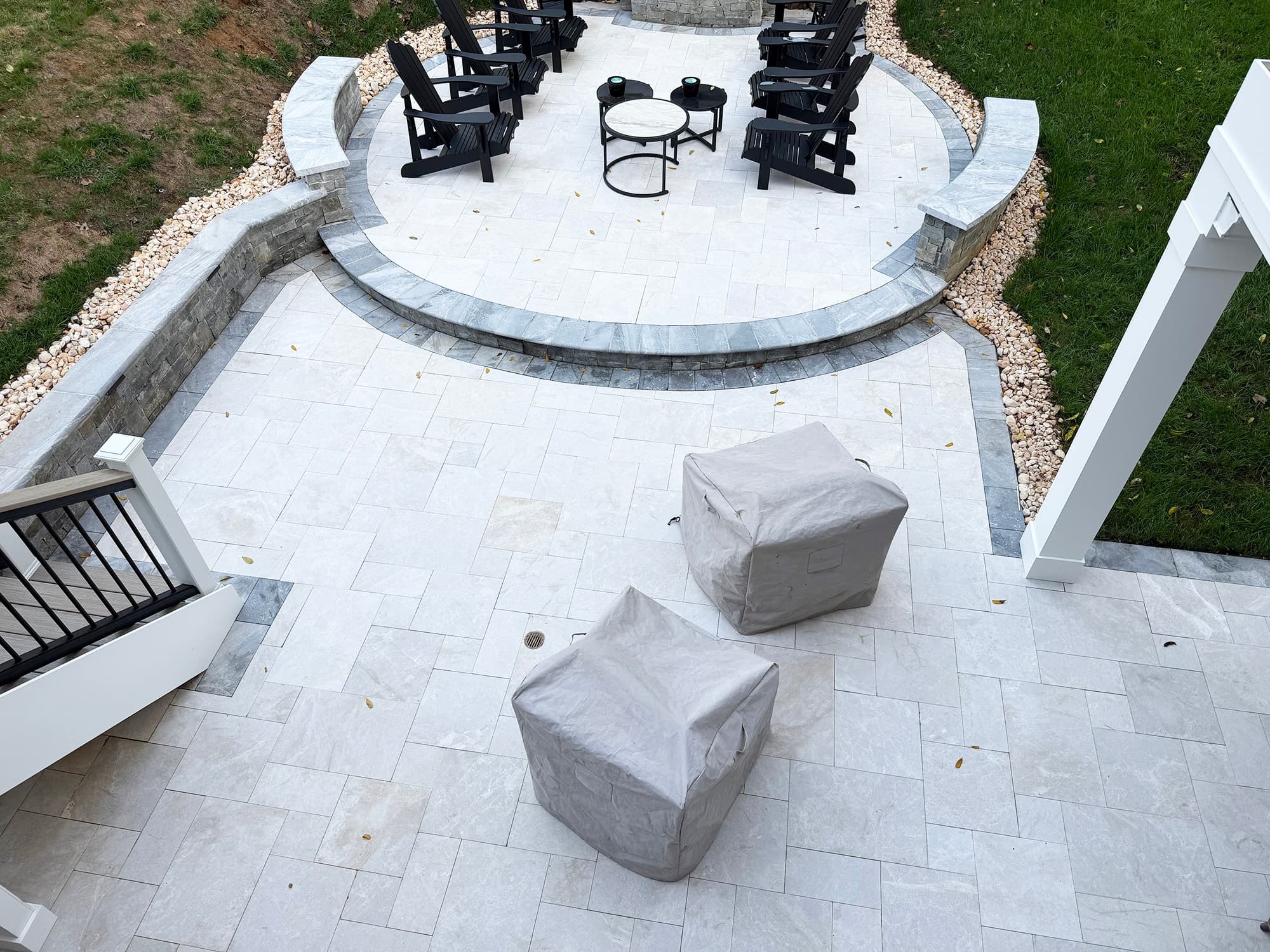 Outdoor patio installation featuring Stonelux Pera Cream Marble Sand Blasted French Pattern 3cm Paver. The elegant marble pavers create a sophisticated look with a sandblasted finish. Surrounded by lush green grass, stone wall, and black Adirondack cha...