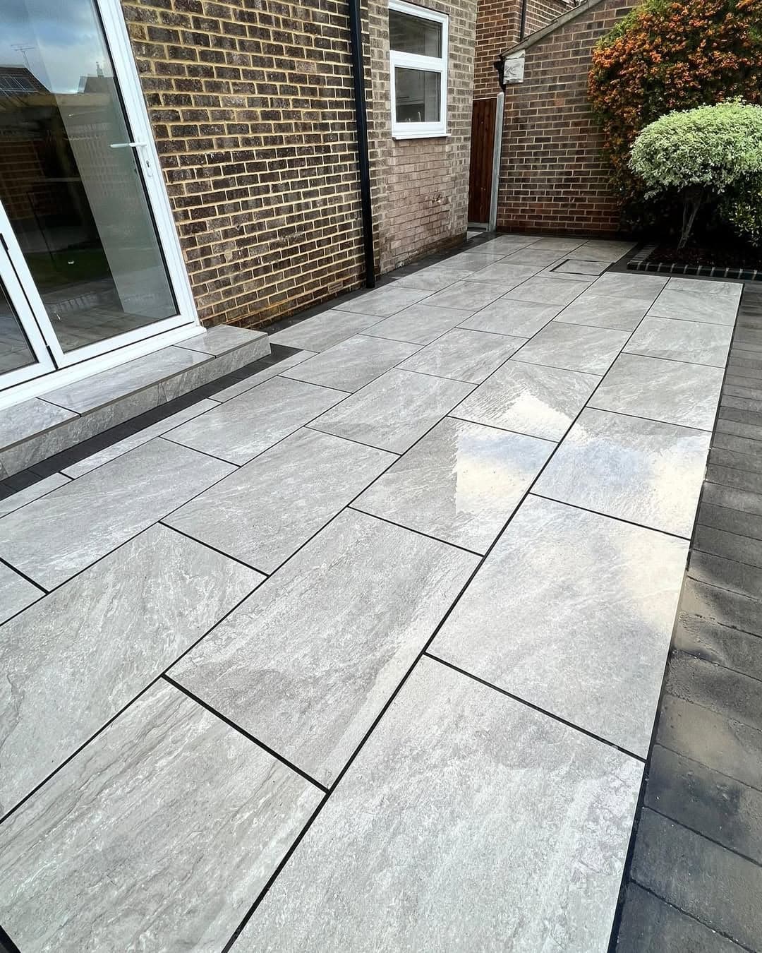 Rasa Gray Matte 24x48x2cm Rectified Porcelain Paver installed as a modern patio. The gray tones and matte finish complement the brick exterior and lush landscaping. Ideal for hardscape projects seeking durable and elegant porcelain solutions.