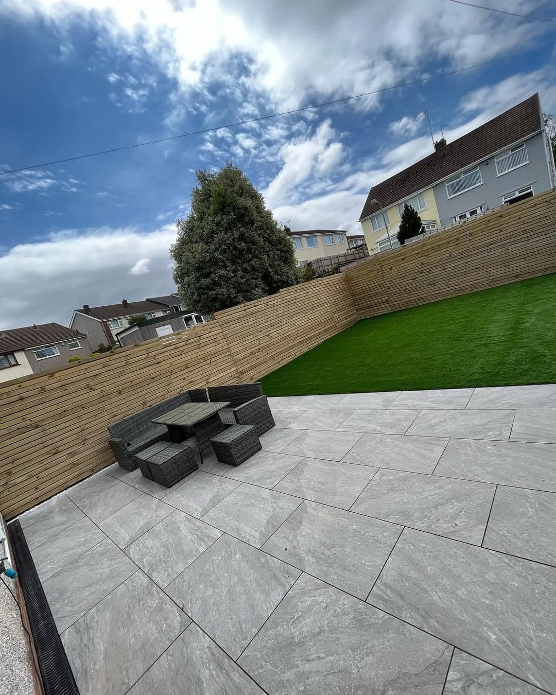 Outdoor patio featuring Rasa Gray Matte 24x48x2cm Rectified Porcelain Pavers by STONELUX. The light gray, matte finish complements a modern garden setting with lush green grass, wooden fencing, and wicker outdoor furniture. Ideal for contemporary hards...