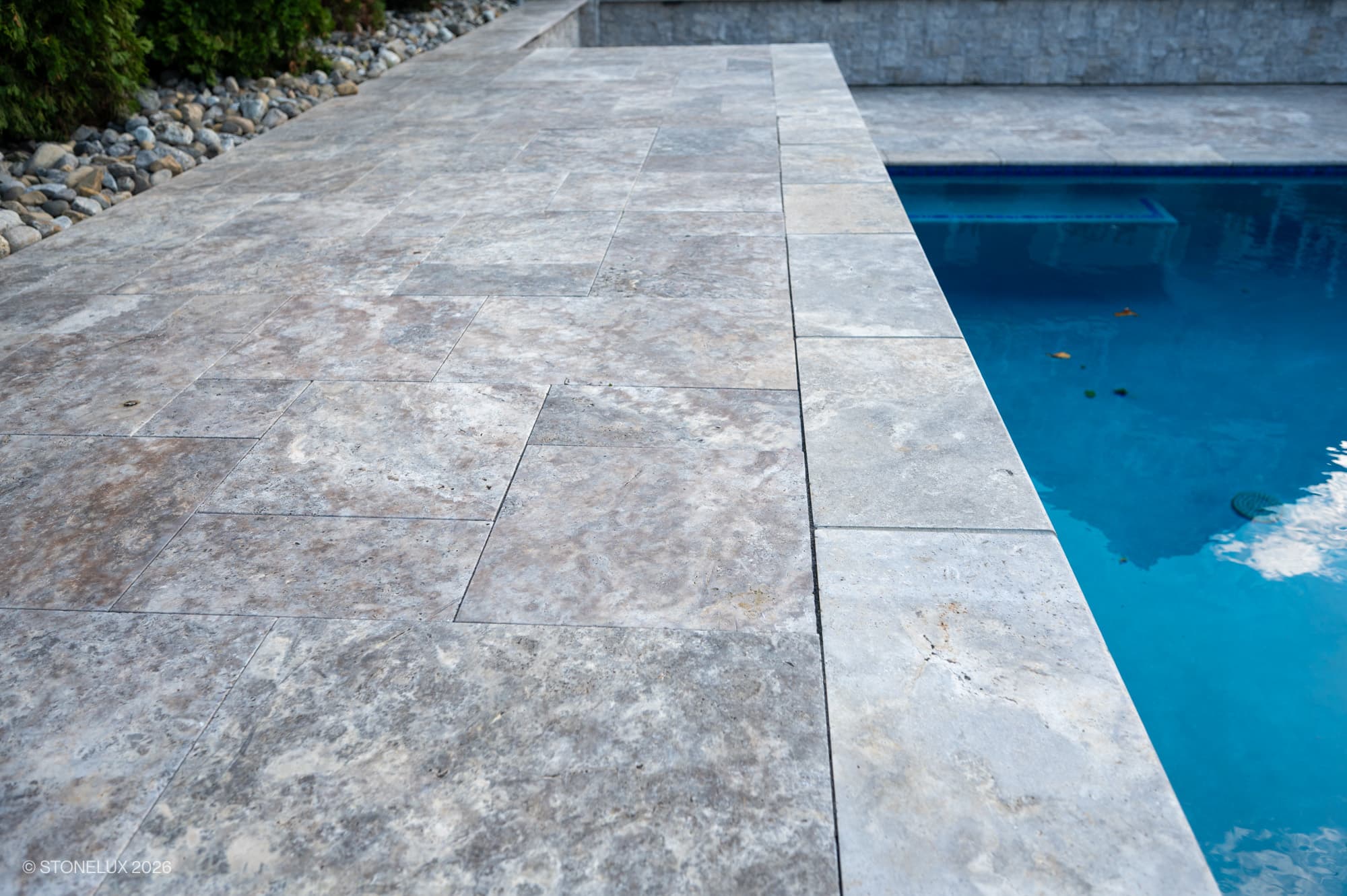 Silver Titanium Travertine Tumbled French Pattern 3cm Paver used as pool coping and deck. The natural stone's gray and silver hues with tumbled finish complement the clear blue pool water, surrounded by stone landscaping, creating a luxurious outdoor s...