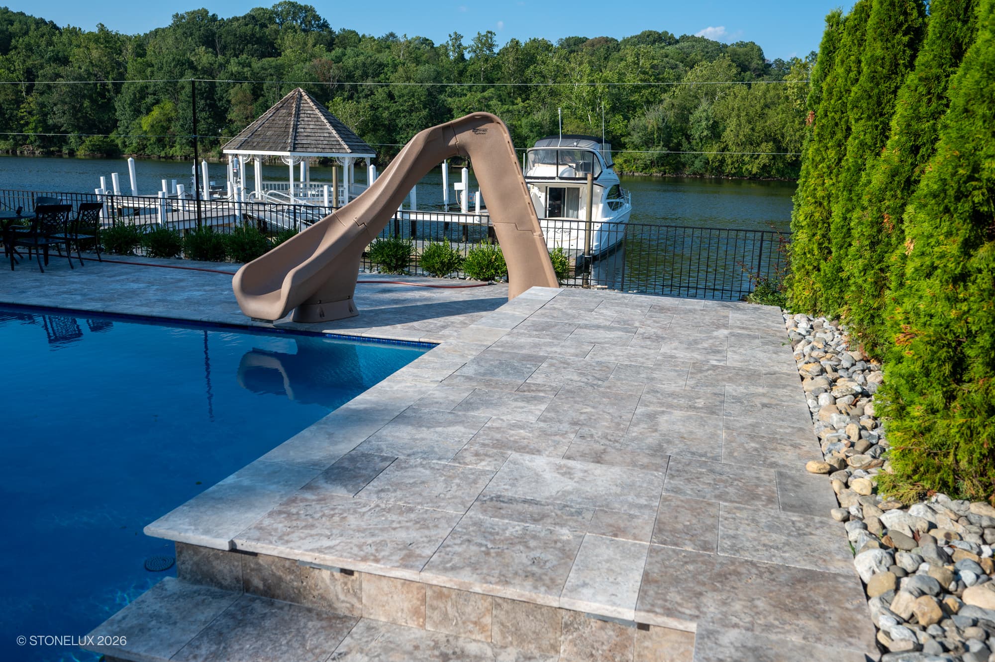 Silver Titanium Travertine Tumbled French Pattern 3cm Paver used for a luxurious pool deck. The natural stone's subtle grey tones complement the sparkling blue pool, beige slide, and lush green landscaping, creating a serene riverside retreat.