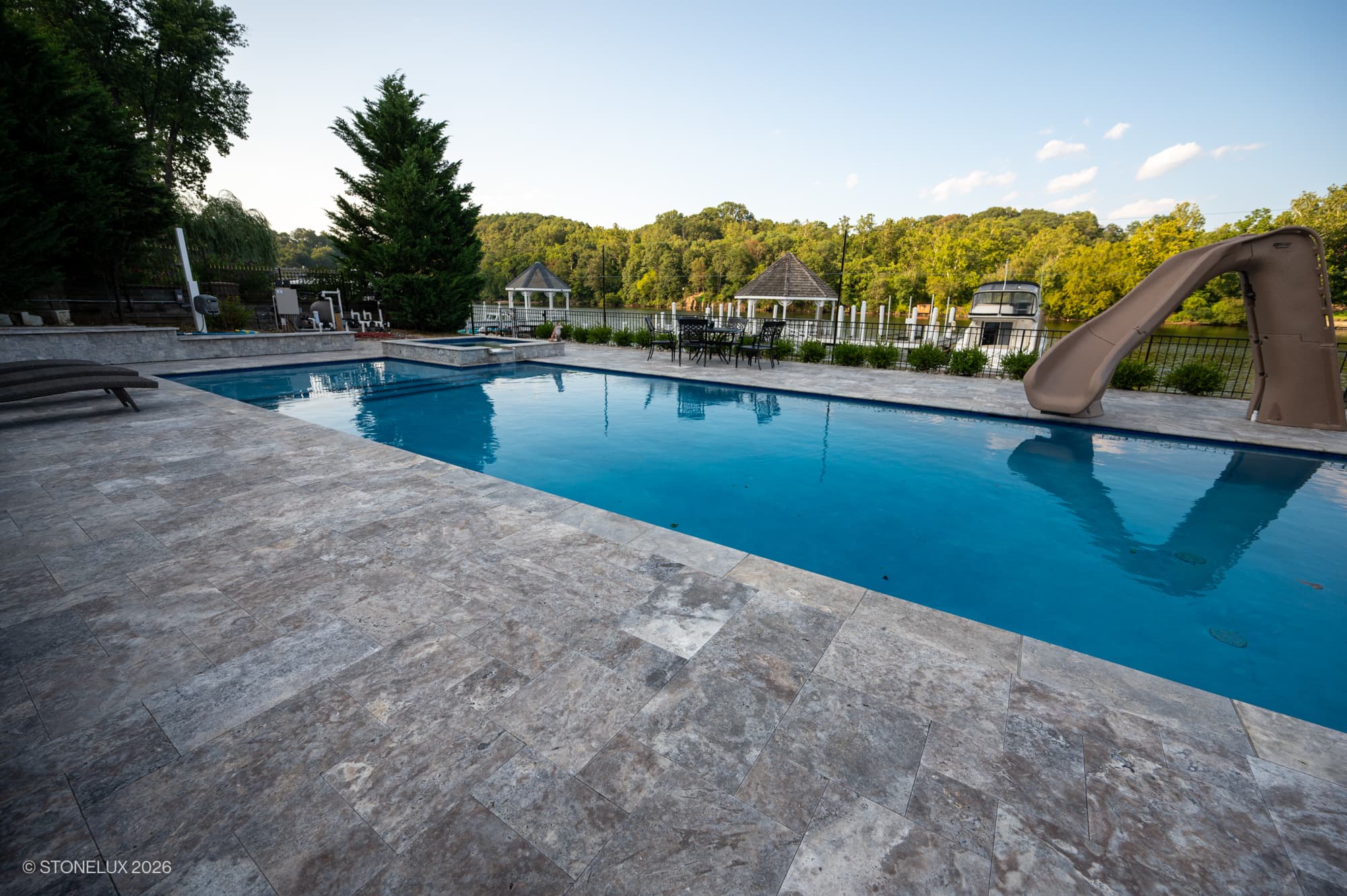 Luxurious poolside installation featuring Silver Titanium Travertine Tumbled French Pattern 3cm Paver by STONELUX. The travertine's natural stone texture and elegant gray tones complement the clear blue pool, surrounded by lush greenery and modern outd...