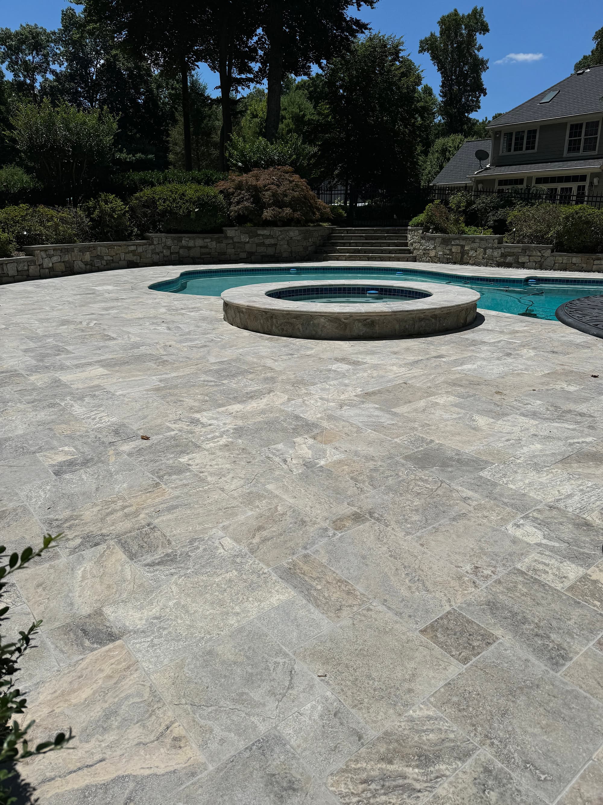 Elegant backyard pool area featuring Silver Travertine Tumbled French Pattern 3cm Paver - Premium by STONELUX. The natural stone pavers, with subtle gray tones and tumbled texture, create a sophisticated patio surrounding a pool with lush greenery and ...