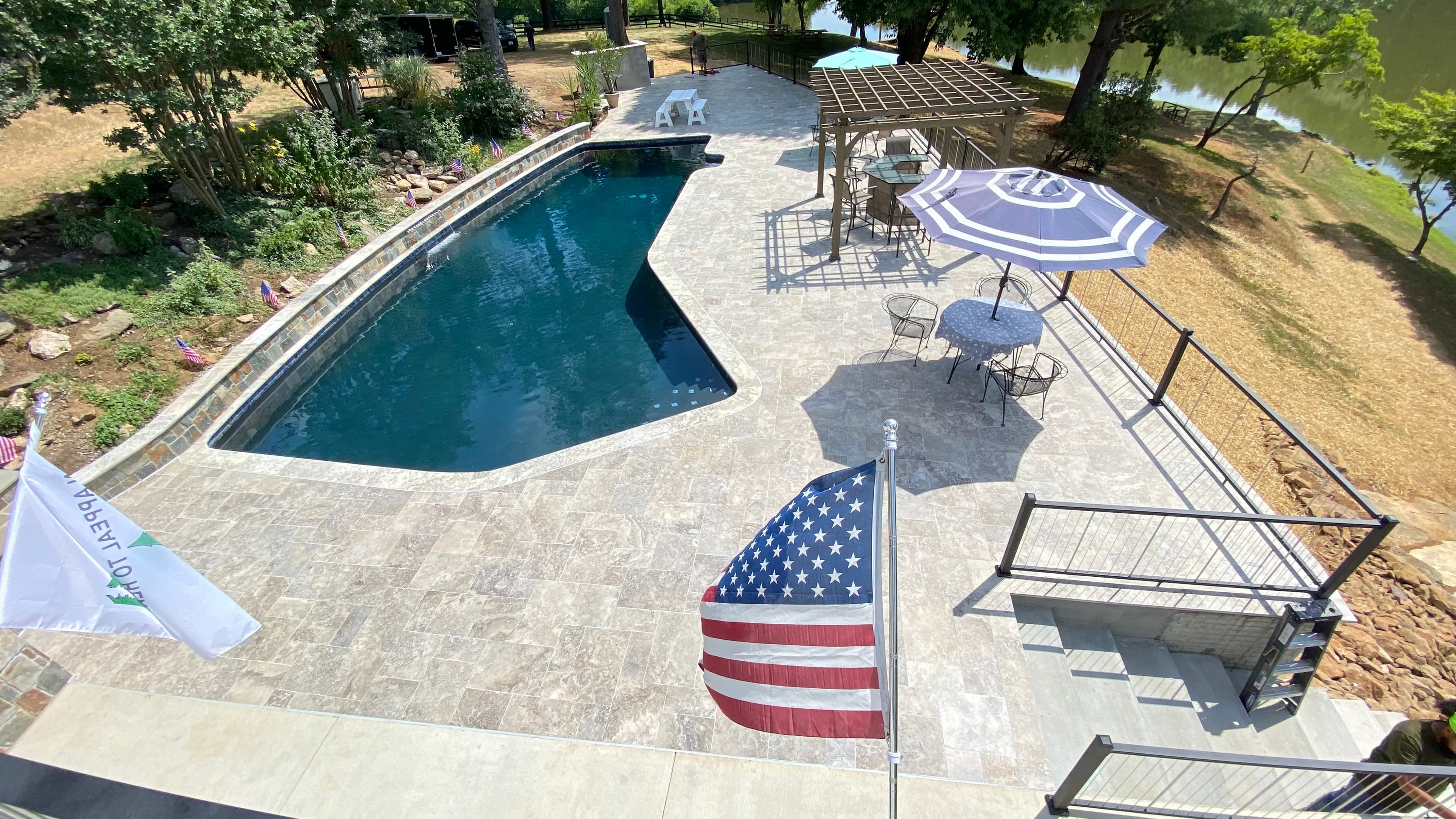 Elevate your outdoor space with Silver Travertine Tumbled French Pattern 3cm Paver by STONELUX. This premium natural stone covers a pool deck, blending seamlessly with a serene blue pool, modern pergola, and lush landscaping for a luxurious hardscape p...