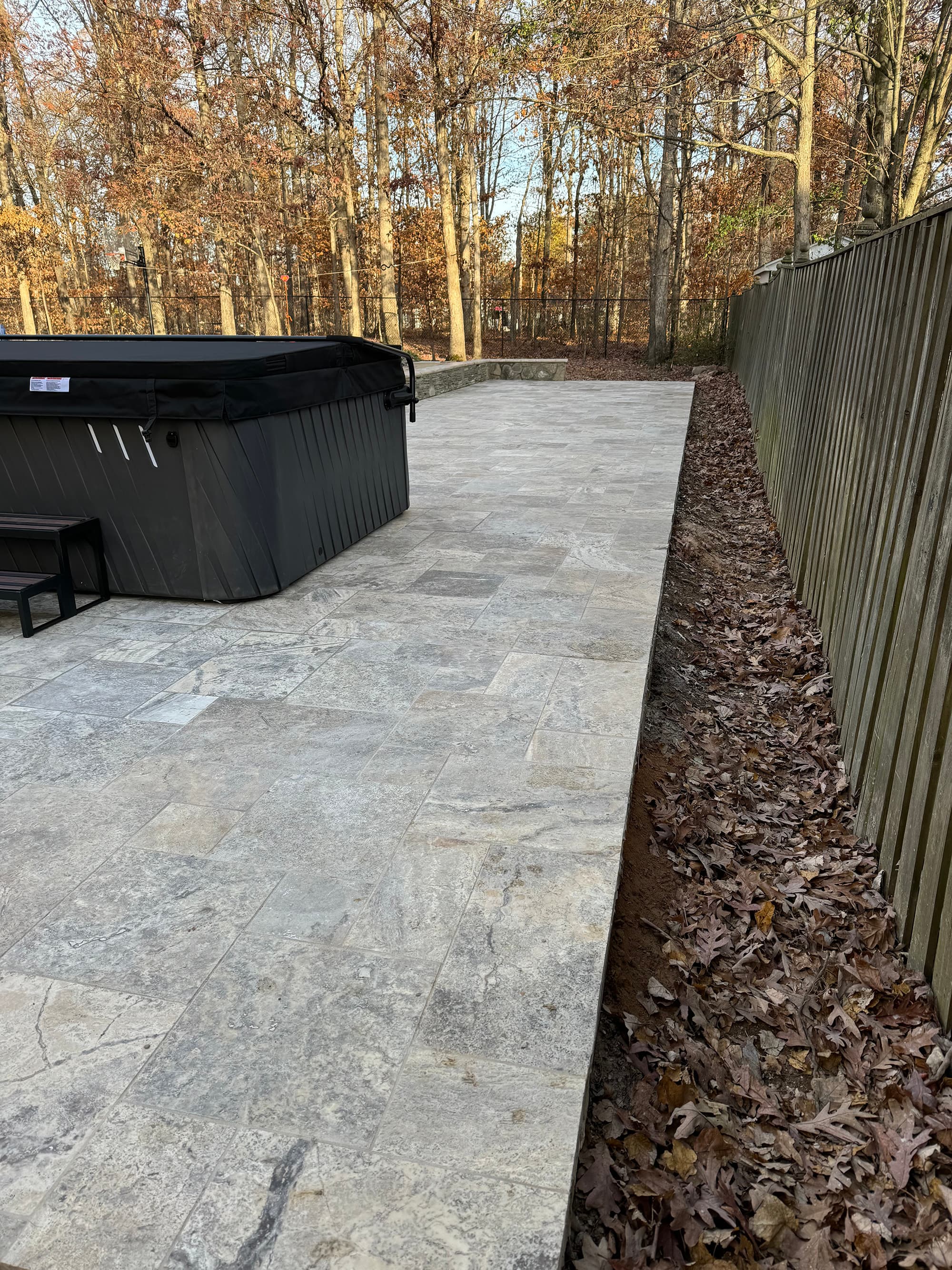 Silver Travertine Tumbled French Pattern 3cm Paver by STONELUX installed as a patio. The natural stone's subtle gray tones and textured finish complement the wooded background and modern hot tub, enhancing the outdoor space with elegance and durability.