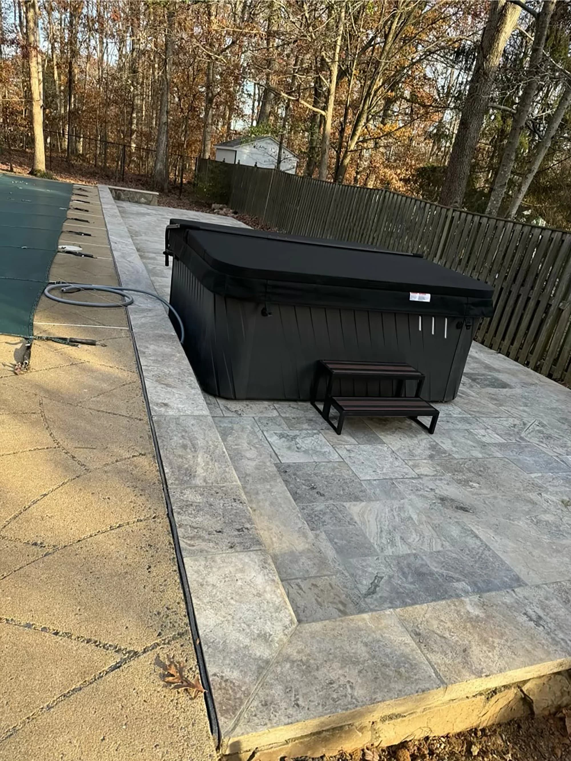 Silver Travertine Tumbled French Pattern 3cm Pavers installed around a hot tub in a backyard setting. The natural stone's soft gray hues and tumbled finish complement a serene landscape with autumn trees and a wooden fence, ideal for outdoor hardscape ...