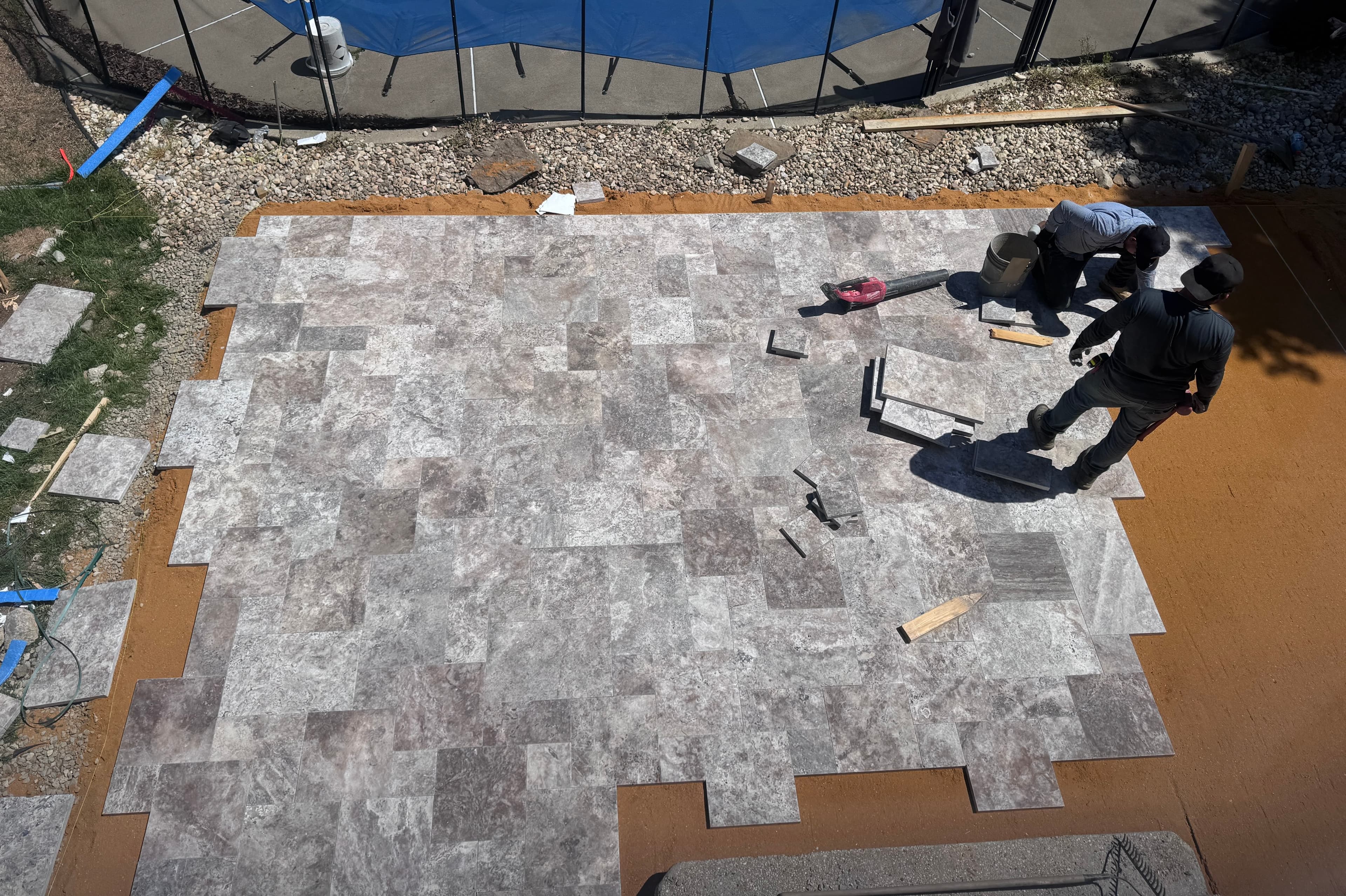 Installation of Silver Titanium Travertine Tumbled French Pattern 3cm Pavers by STONELUX. Workers lay natural stone pavers around a pool area, bordered with gravel and grass. The tumbled finish and varied gray tones create a sophisticated, durable outd...