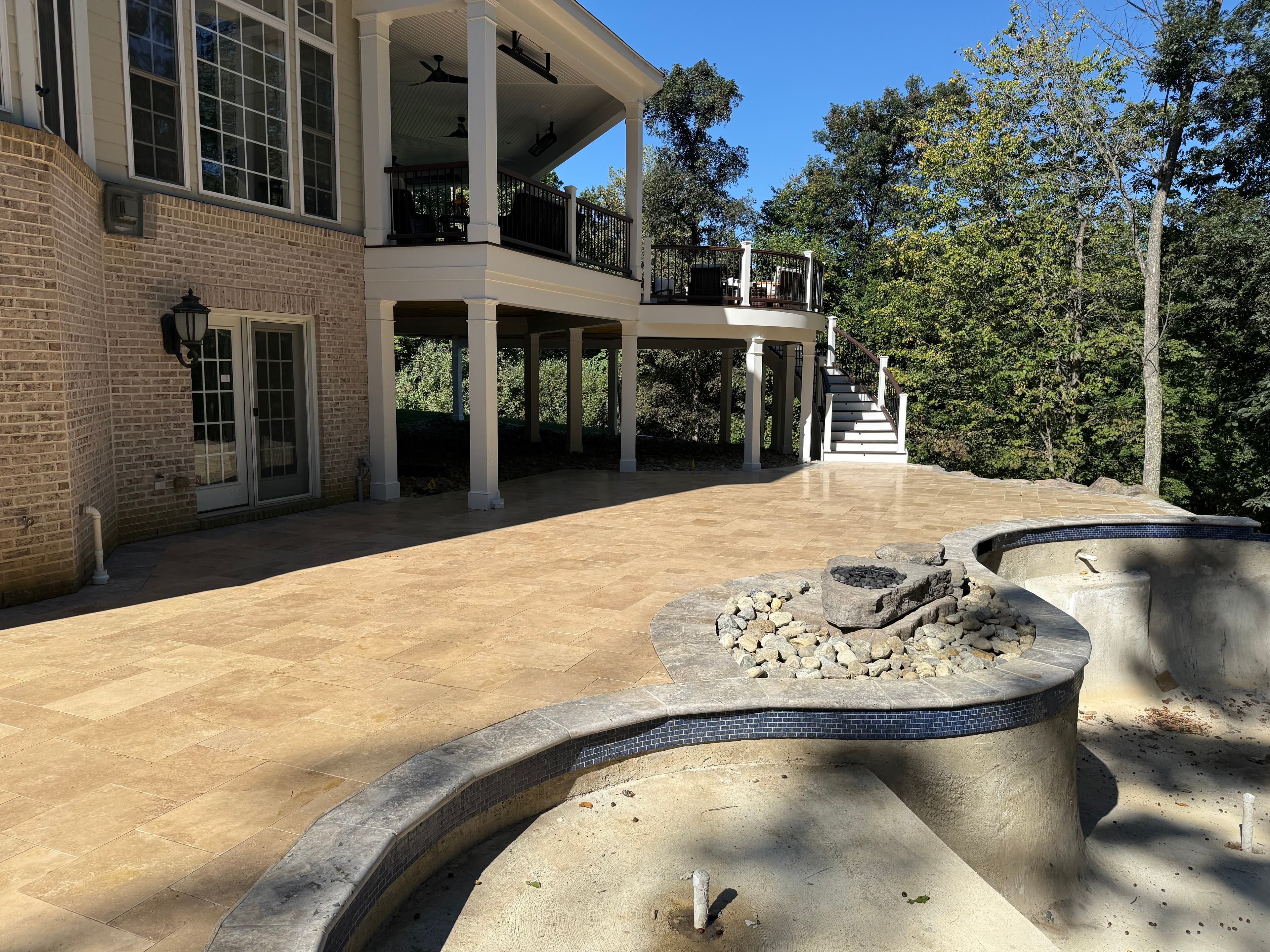 Spacious patio featuring Walnut Travertine Tumbled French Pattern 3cm Paver - Premium, showcasing natural stone elegance. Complemented by a traditional brick house, lush greenery, and an unfinished pool area, ideal for luxurious outdoor hardscape proje...
