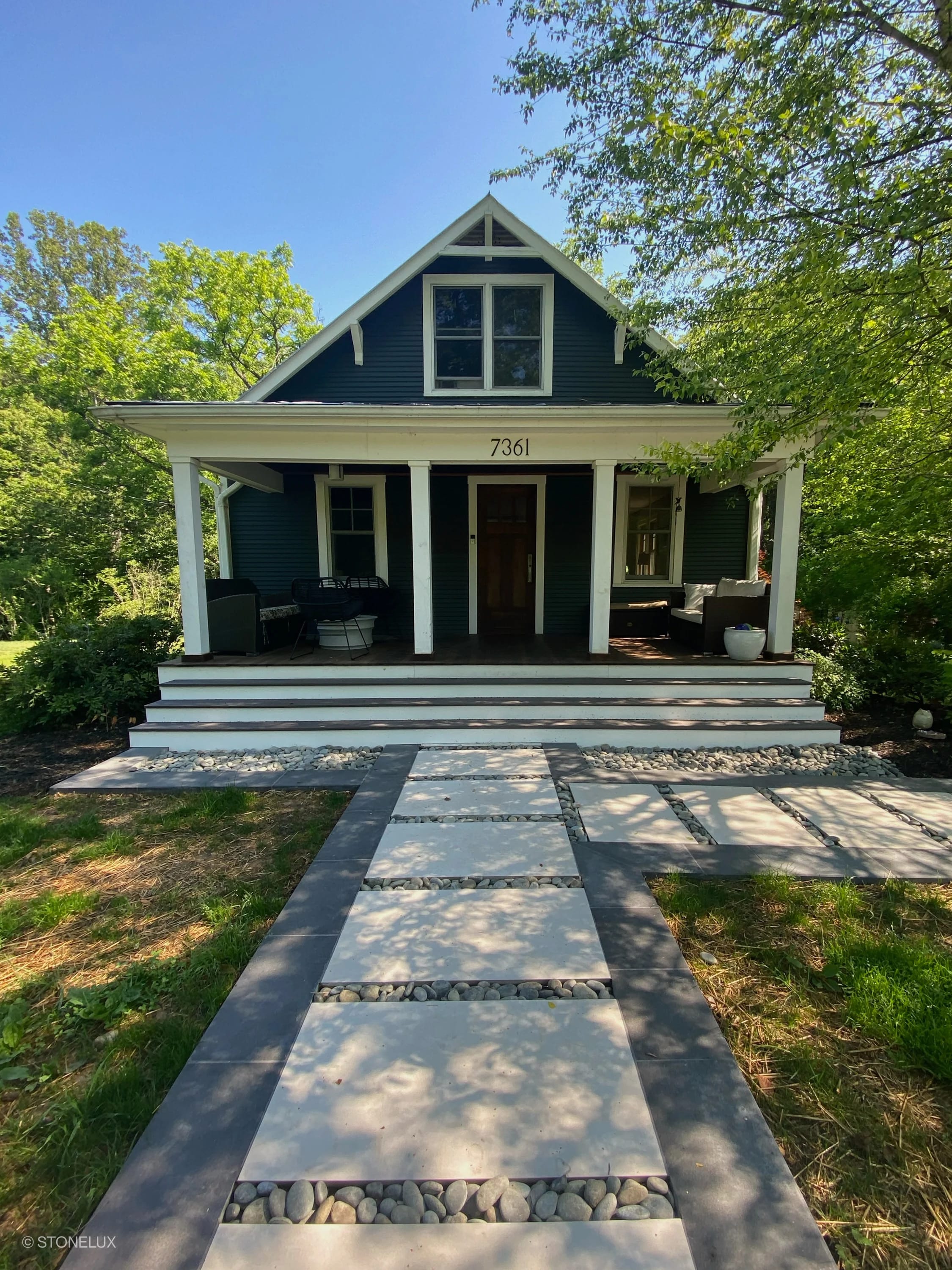 Front entrance showcasing Overland Pearl Matte 24x48x2cm Rectified Porcelain Paver path leading to a traditional dark-painted house. The pavers are interspersed with decorative stones, surrounded by lush landscaping, creating a modern yet classic aesth...