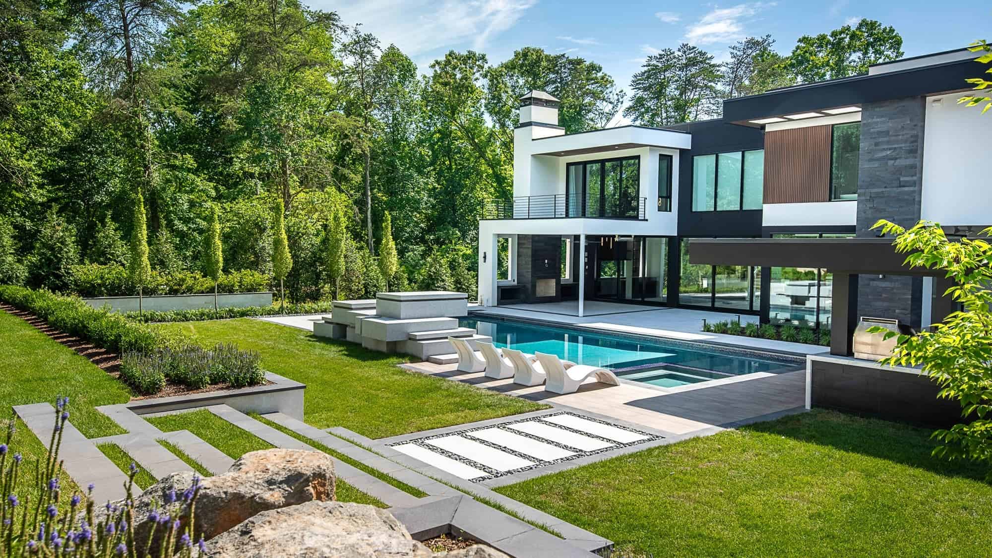 Transform your outdoor space with STONELUX Porcelain Pavers—ideal for sunny days by the pool, hot tub, or landscaped garden beds. Enjoy a modern home with large windows, balcony, stone pathways, and a manicured lawn amid lush trees.