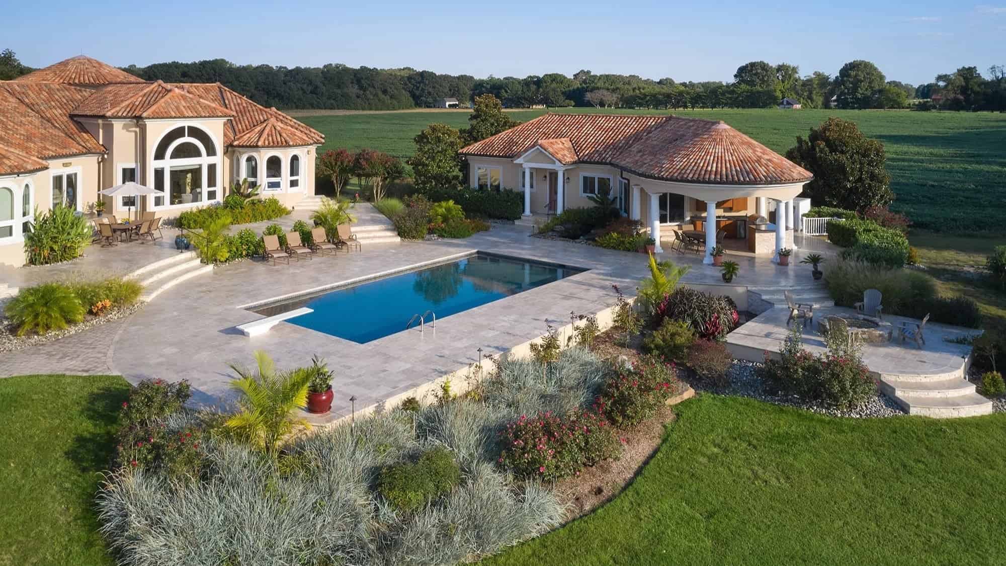 Aerial view of a luxurious house with a terracotta roof, large pool bordered by STONELUX Travertine Pavers, landscaped gardens, lounge chairs, covered patio, and green lawns surrounded by open fields.