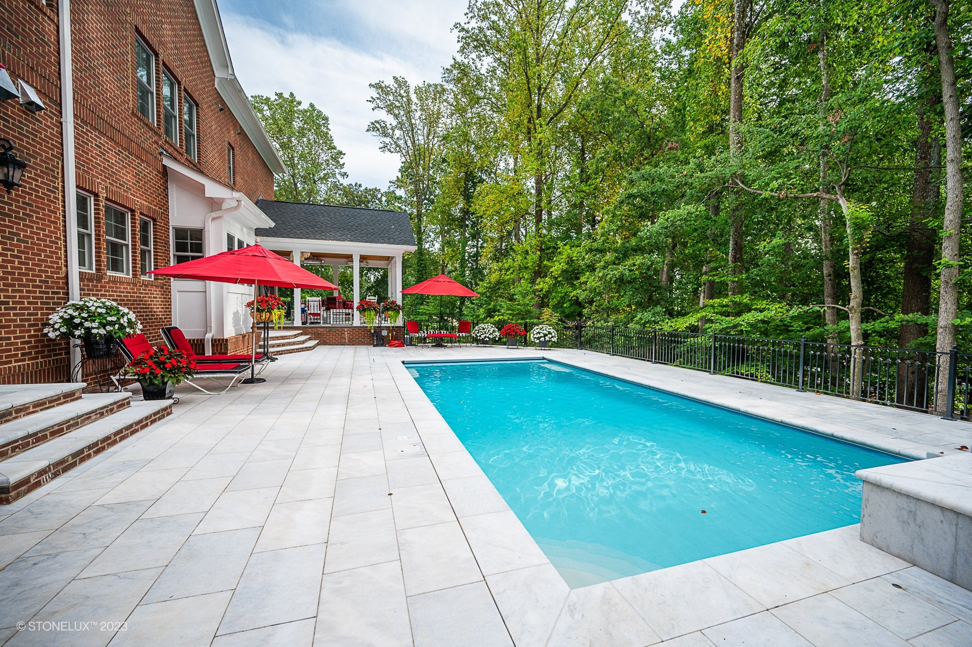 Afyon White Marble pool deck installation by STONELUX. Elegant natural stone pavers surround a vibrant blue pool, complemented by red umbrellas and lounge chairs. Brick home backdrop and lush green trees create a luxurious outdoor setting.
