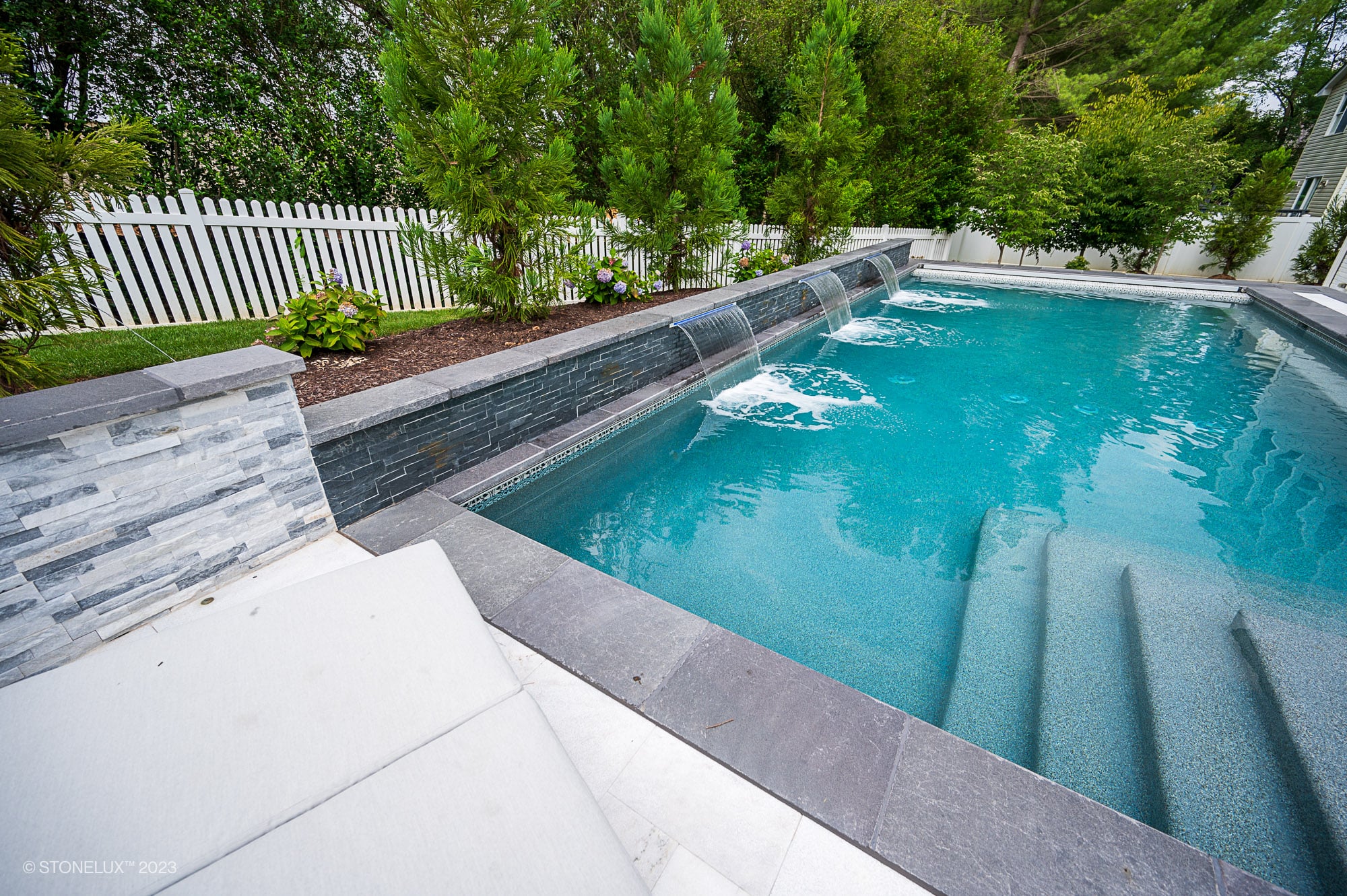 Luxurious pool area featuring STONELUX Black Basalt natural stone as pool coping and wall accents. Crystal-clear water, cascading waterfalls, and lush greenery complement the modern design. Perfect for hardscape projects and elegant pool renovations.