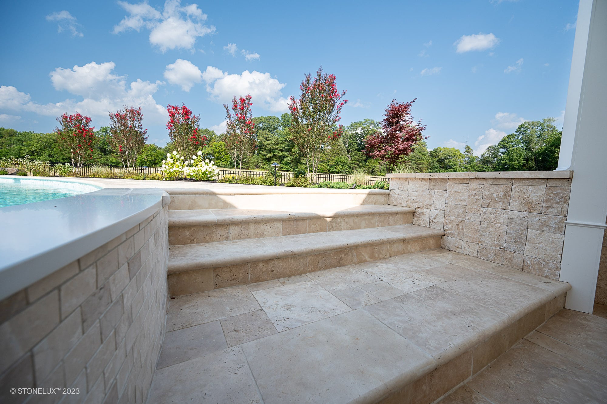 Ivory Travertine natural stone steps and pool coping create a seamless transition between the pool area and garden. The light beige tones and subtle veining enhance the outdoor landscape with vibrant red and green foliage, offering a serene, elegant look.