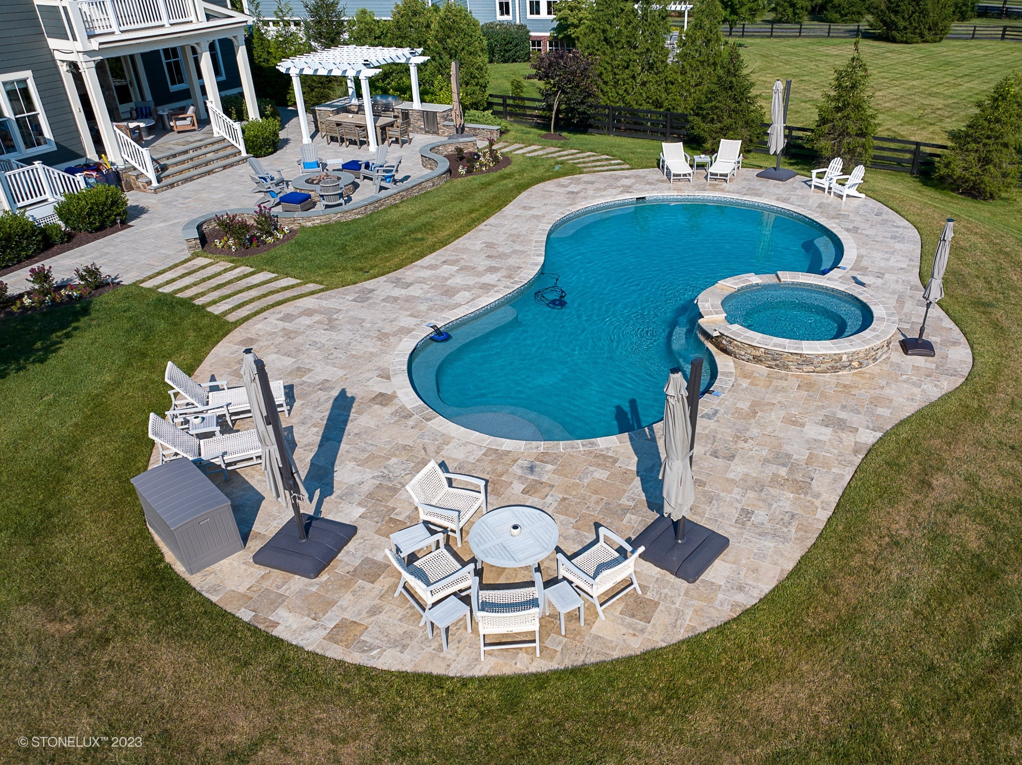 Luxurious backyard installation featuring Philly Travertine natural stone pavers surrounding a curvaceous pool and spa. The patio complements the modern outdoor furniture, pergola, and lush landscaping, creating a serene, elegant outdoor living space.