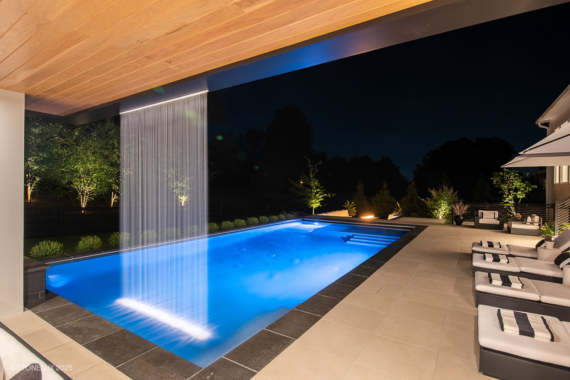 Luxurious outdoor pool area featuring STONELUX Porcelain Paver in natural stone design. Elegant pool deck with modern water feature, ambient lighting, and stylish lounge furniture. Complemented by lush landscaping and serene night ambiance. Ideal for p...