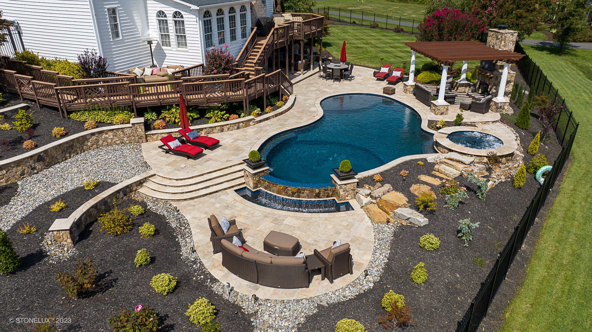Luxurious backyard installation featuring Walnut Travertine pavers by STONELUX, used for an elegant patio and pool deck. The design includes a custom-shaped pool, spa, wooden pergola, outdoor seating, vibrant landscaping, and a cascading water feature,...