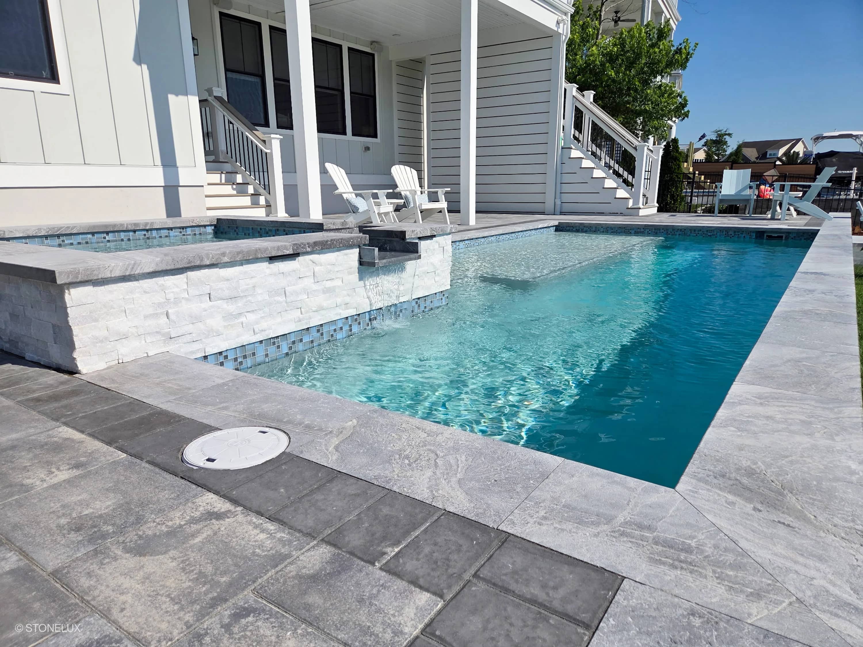 Modern pool design featuring Aegean Grey Marble Sand Blasted 16x24x2 Eased Edge Coping. The marble coping elegantly frames the pool, complementing the light gray porcelain pavers. Crisp blue water reflects the contemporary architecture and lush landsca...