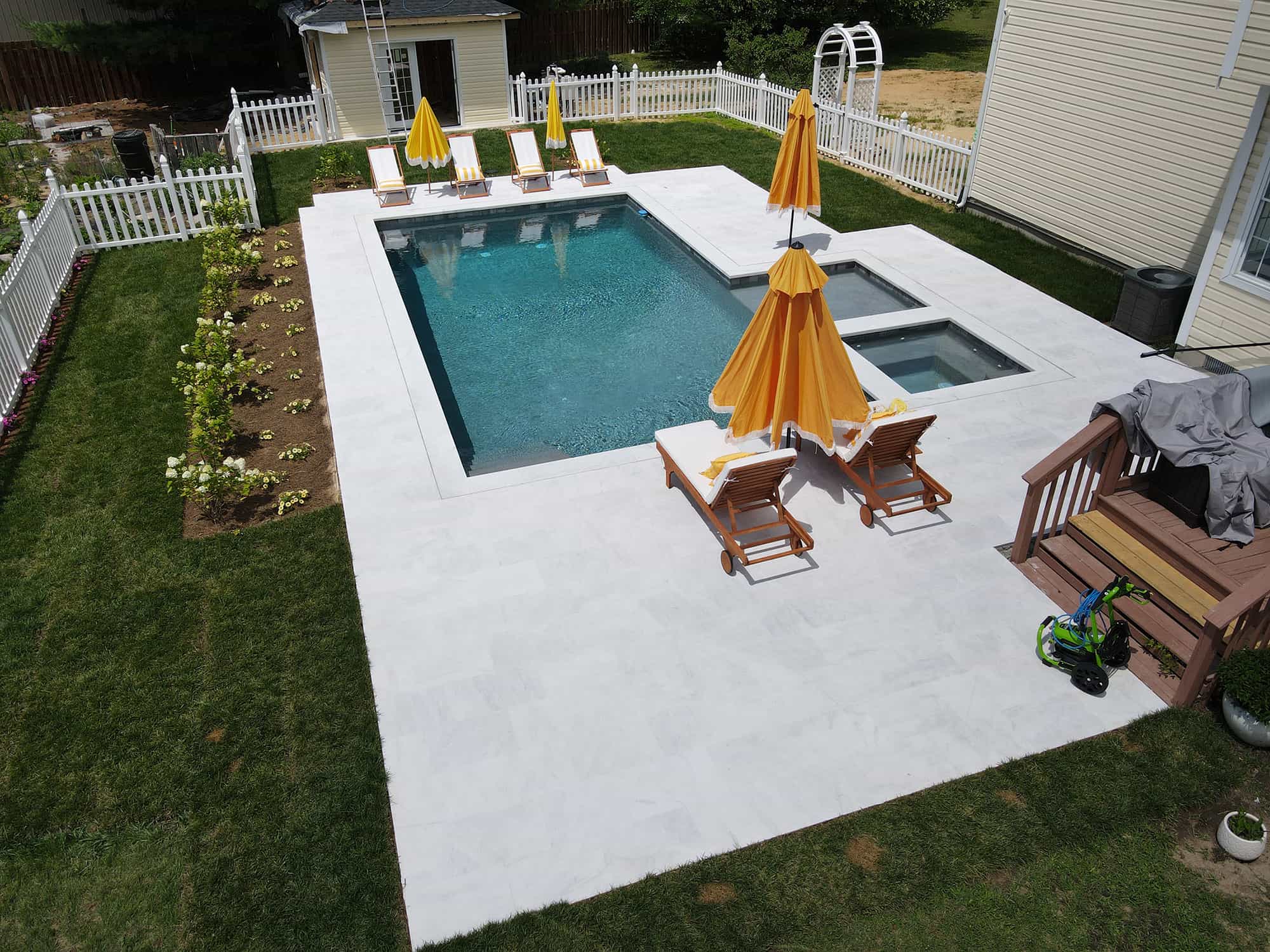 Afyon White Marble Sand Blasted 16x24 3cm Paver installation at a poolside. The marble's light hue and sandblasted texture complement the clear blue pool, surrounded by lush greenery, white fencing, and wooden lounge chairs with vibrant orange umbrella...