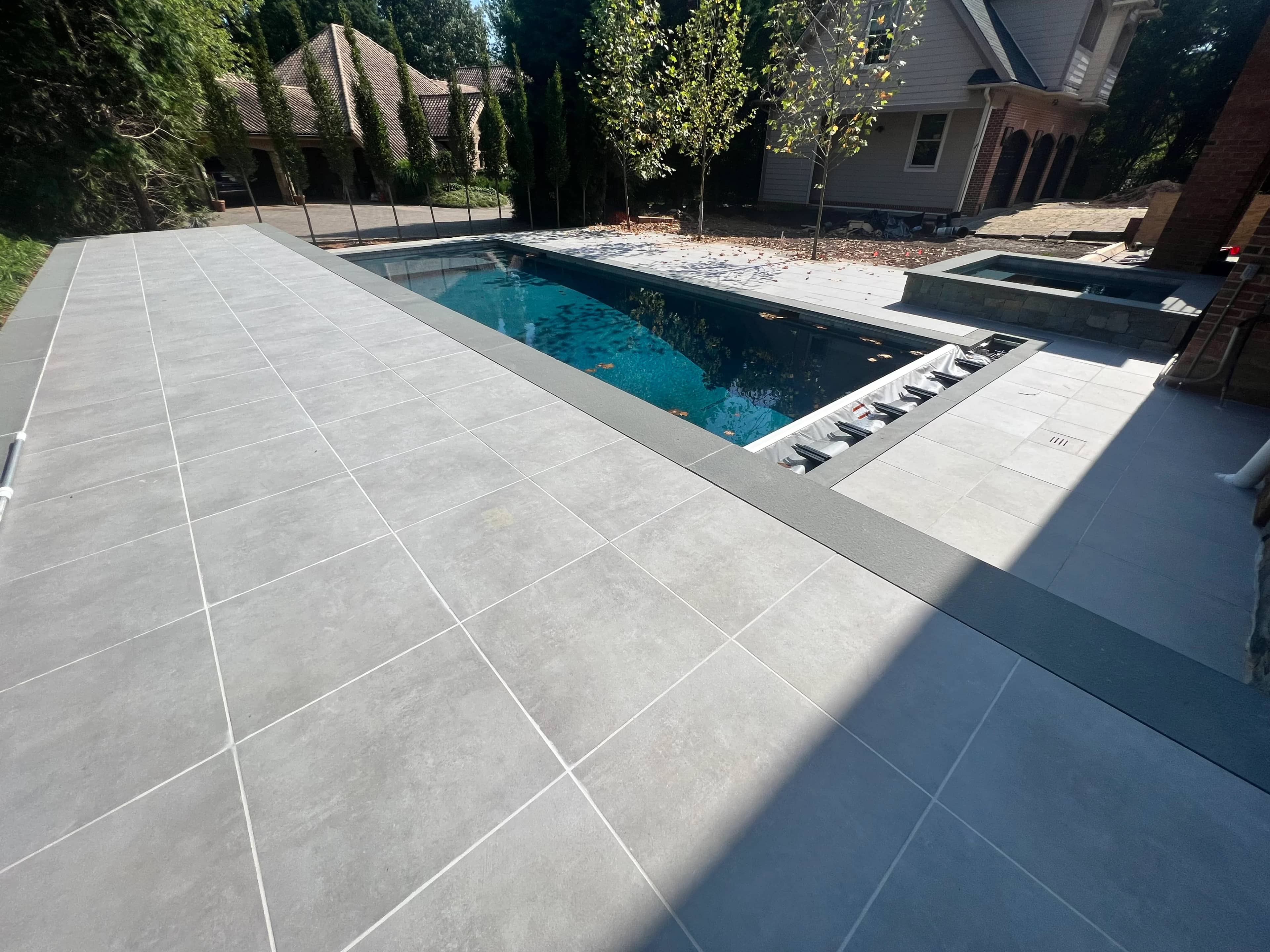 Premium outdoor pool deck featuring Ark Silver Matte 24x24x2cm Rectified Porcelain Paver from Stonelux. The sleek, matte finish complements the clear blue pool water and surrounding trees, creating a modern, elegant hardscape design. Ideal for luxury r...