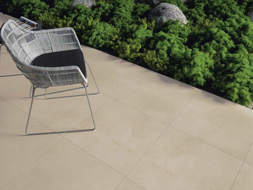 Luna Sand Matte 24x24x2cm Rectified Porcelain Paver installed as patio flooring. The pavers feature a smooth matte finish with subtle sand tones. Paired with modern outdoor wicker chairs, lush green landscaping, and natural stone accents for a contempo...