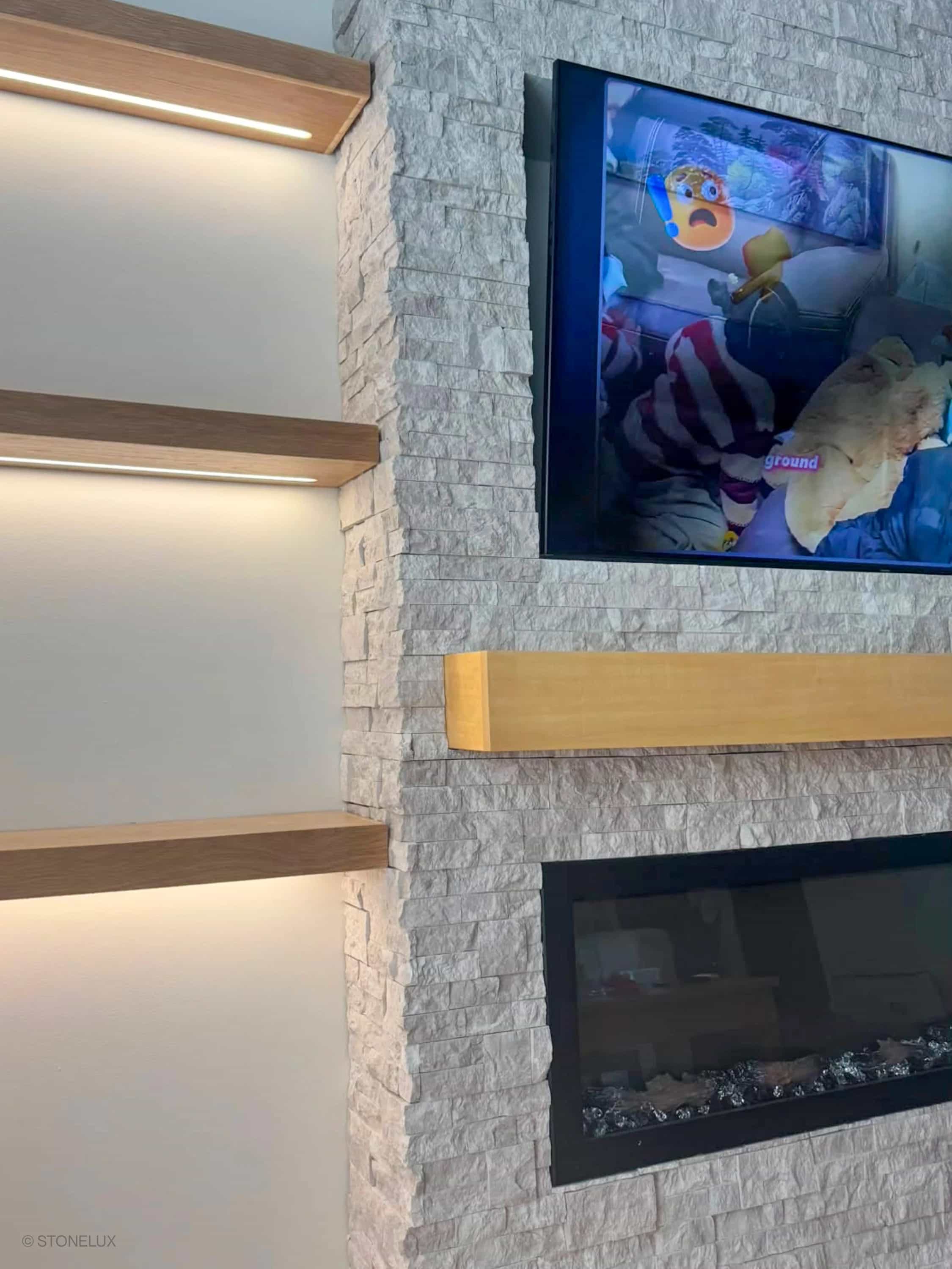 Pera Cream Marble Splitface Ledger Panel - 6x24 installed as a modern fireplace surround. The textured marble veneer complements wooden shelves and a sleek TV setup, creating a contemporary living space with warm, neutral tones and elegant design eleme...