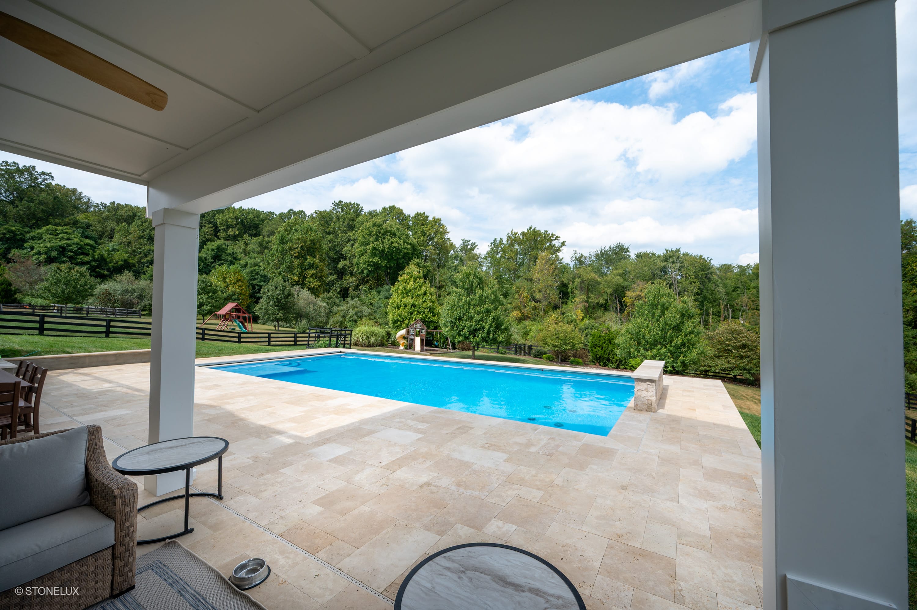 Ivory Travertine Tumbled French Pattern 3cm Paver by STONELUX installed around a luxurious pool. The creamy travertine pavers complement the serene blue water, surrounded by lush greenery, creating a harmonious outdoor oasis ideal for hardscape projects.