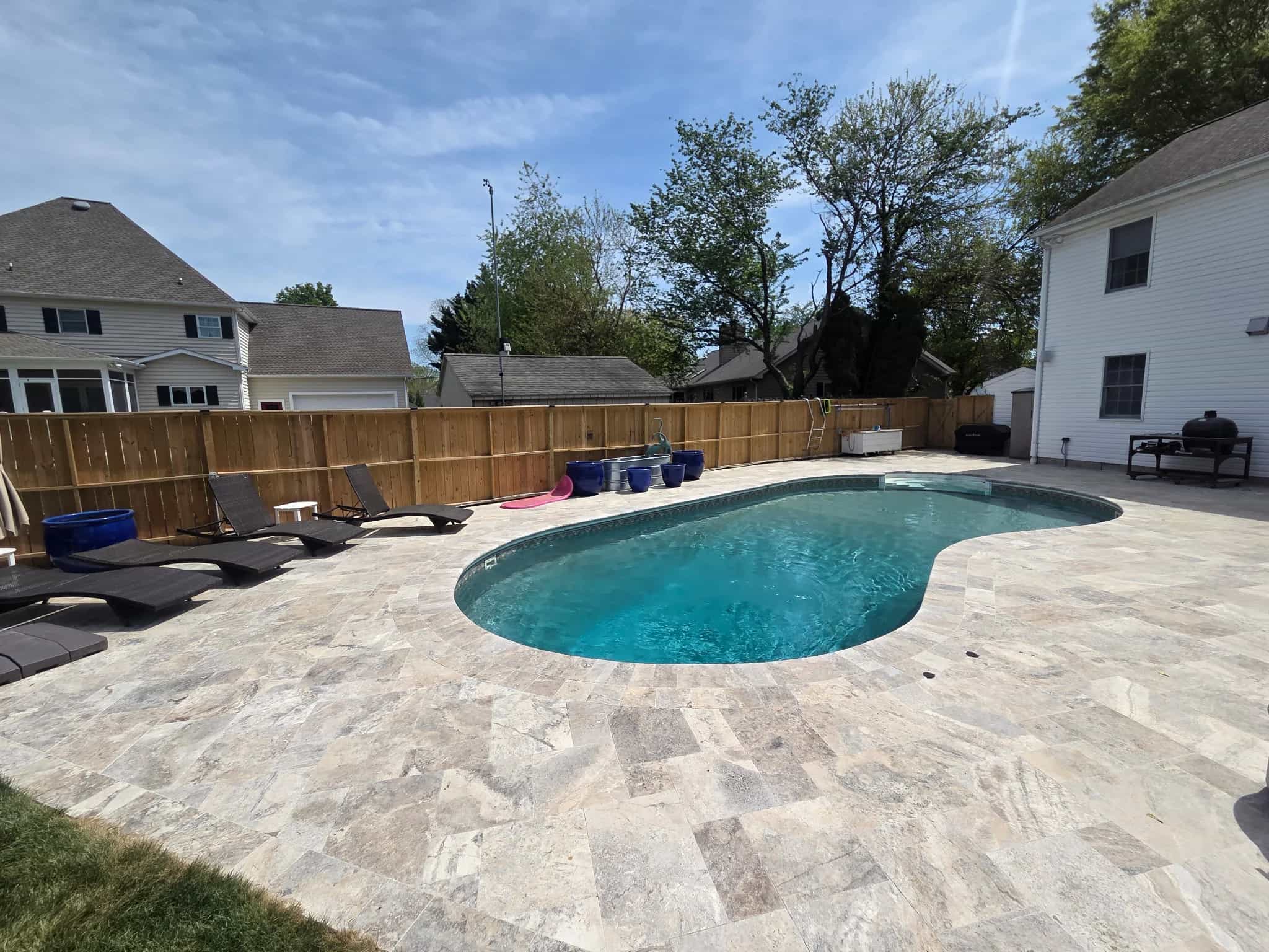Backyard pool deck featuring Silver Travertine Tumbled 16x24 3cm Paver - Select by STONELUX. The natural stone pavers, in soft silver hues with a tumbled finish, elegantly surround a sparkling blue pool. Modern outdoor furniture and a wooden fence comp...