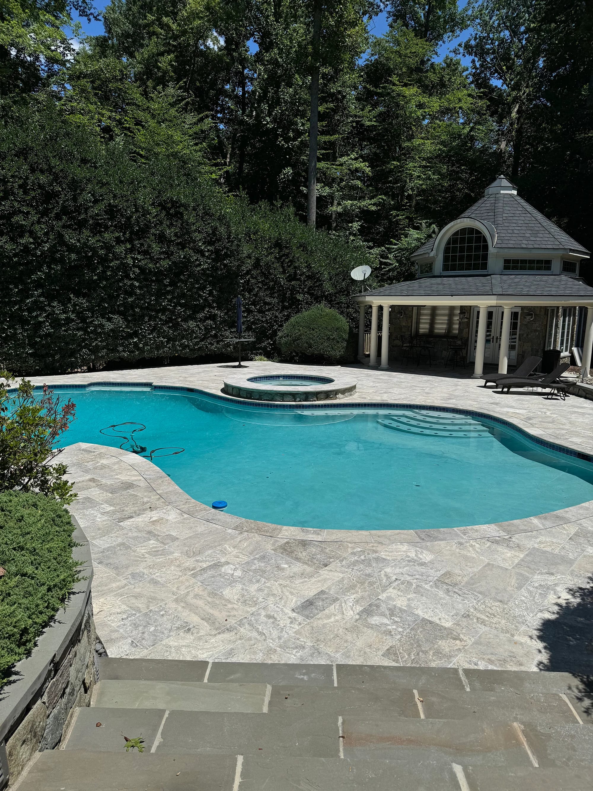Luxurious poolside featuring Silver Travertine Tumbled French Pattern 3cm Paver by STONELUX. The natural stone pavers create a sophisticated patio with their smooth, tumbled finish. Surrounded by lush greenery, a classic gazebo, and sparkling blue pool...