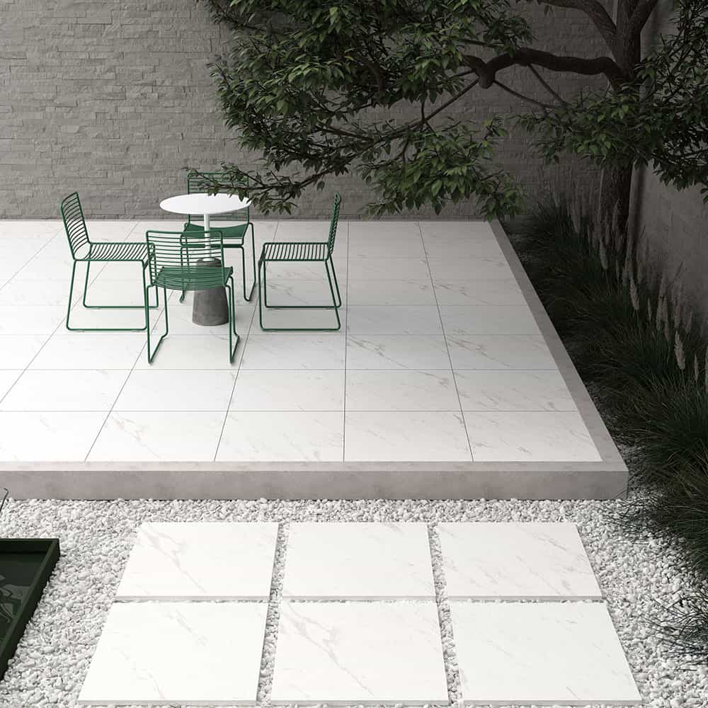 Outdoor patio featuring Tokyo White Matte 24x24x2cm Rectified Porcelain Pavers. The smooth, matte finish provides a modern look, complemented by green metal furniture and lush landscaping. Ideal for contemporary hardscape projects and poolside areas.