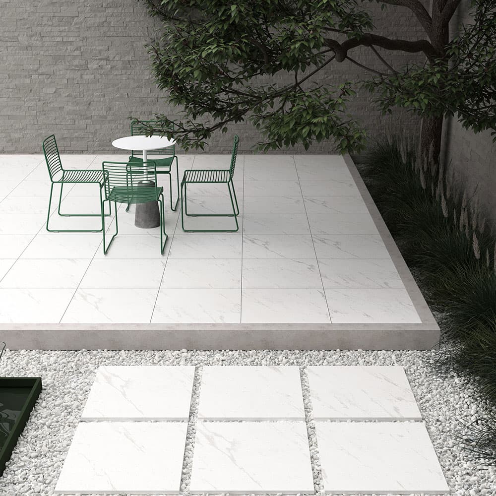 Outdoor patio featuring Tokyo White Matte 24x24x2cm Rectified Porcelain Pavers. The smooth, matte finish provides a modern look, complemented by green metal furniture and lush landscaping. Ideal for contemporary hardscape projects and poolside areas.