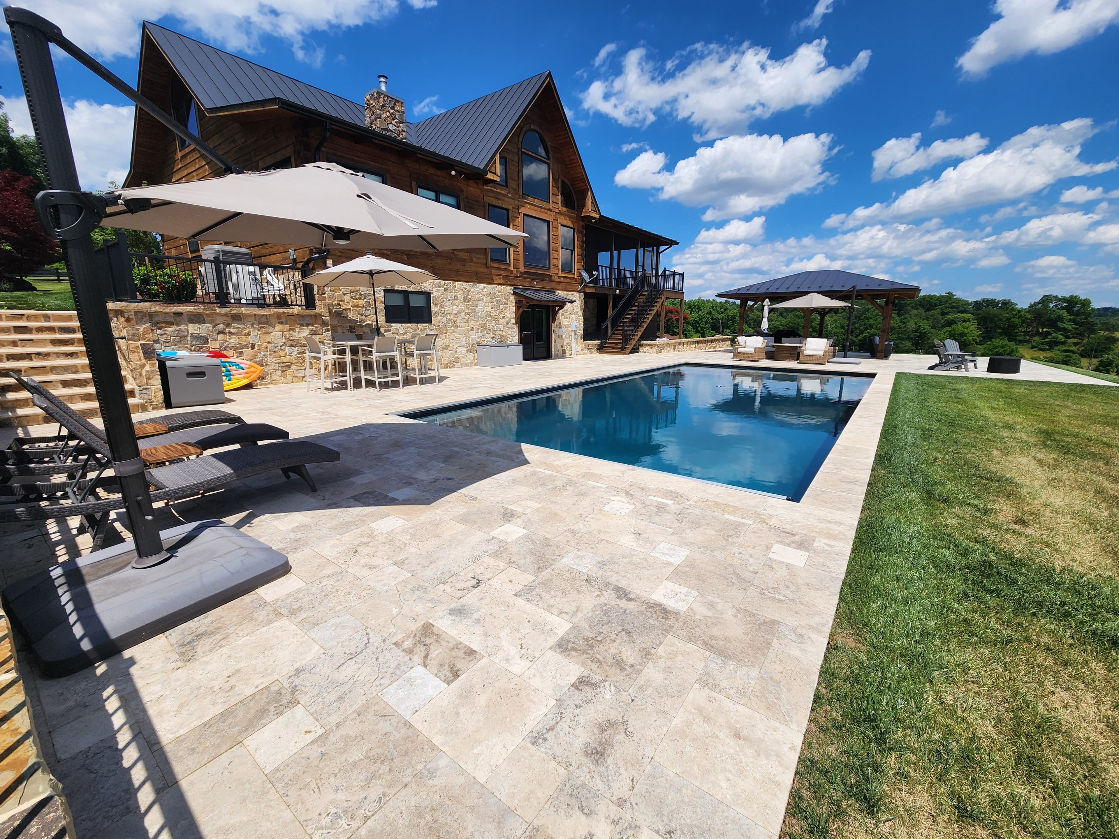 A modern stone patio in Winchester, VA showcases a rectangular pool—this Pool & Patio Project boasts Philly Travertine Elegance, with lounge chairs, umbrellas, and a wooden house among green landscapes beneath a blue sky.