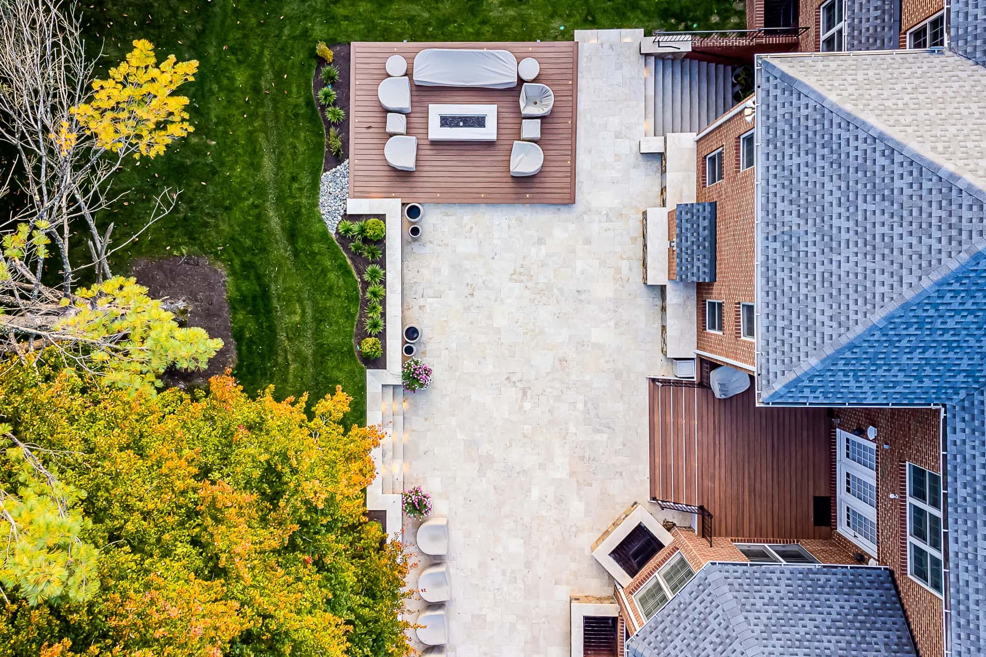 Aerial view of a spacious backyard patio, featuring Ivory Twilight: A Travertine Patio Project with outdoor seating, a fire pit on a wooden deck, stone flooring, green lawn, autumn trees, and part of a large brick house with gray roofing.