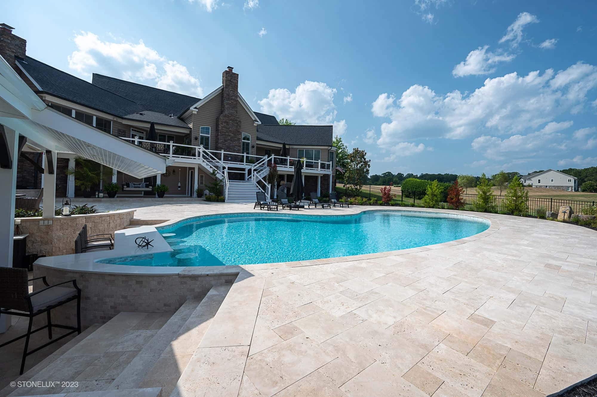 A large, modern backyard features a curved swimming pool and hot tub set in Beige Bliss: Luxurious Travertine. A two-story house with a balcony overlooks the pool, green lawn, and lounge chairs beneath a blue sky with clouds.