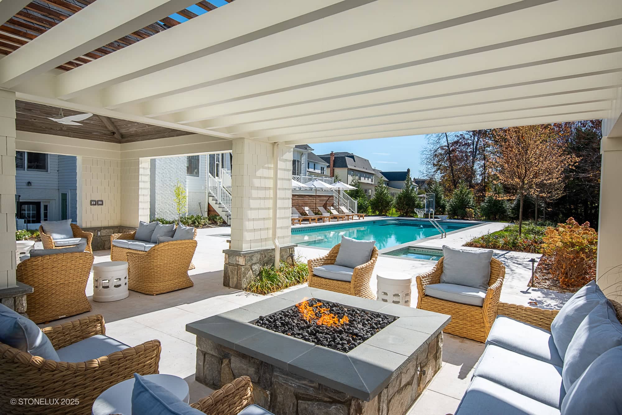 Spacious outdoor patio with wicker seating, a fire pit, and a view of a rectangular swimming pool, surrounded by trees and modern houses—perfect for Ashburn Family Fun and elegant outdoor living in Pera Cream Marble on a sunny day.