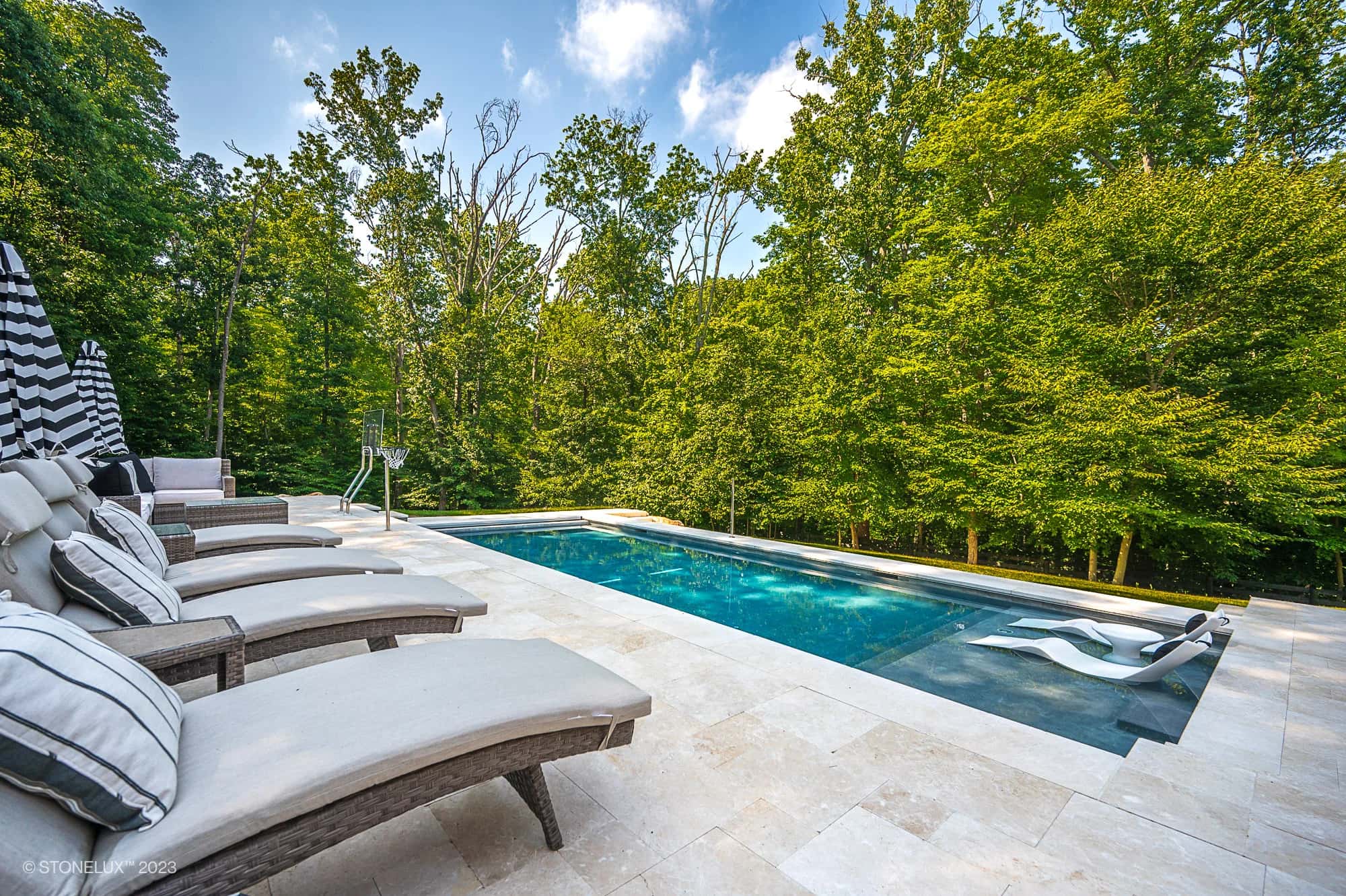 A luxurious outdoor pool with clear blue water, surrounded by trees. Pera Cream Marble tiles line the poolside, where cushioned lounge chairs and striped umbrellas create a relaxing and inviting atmosphere among the trees.