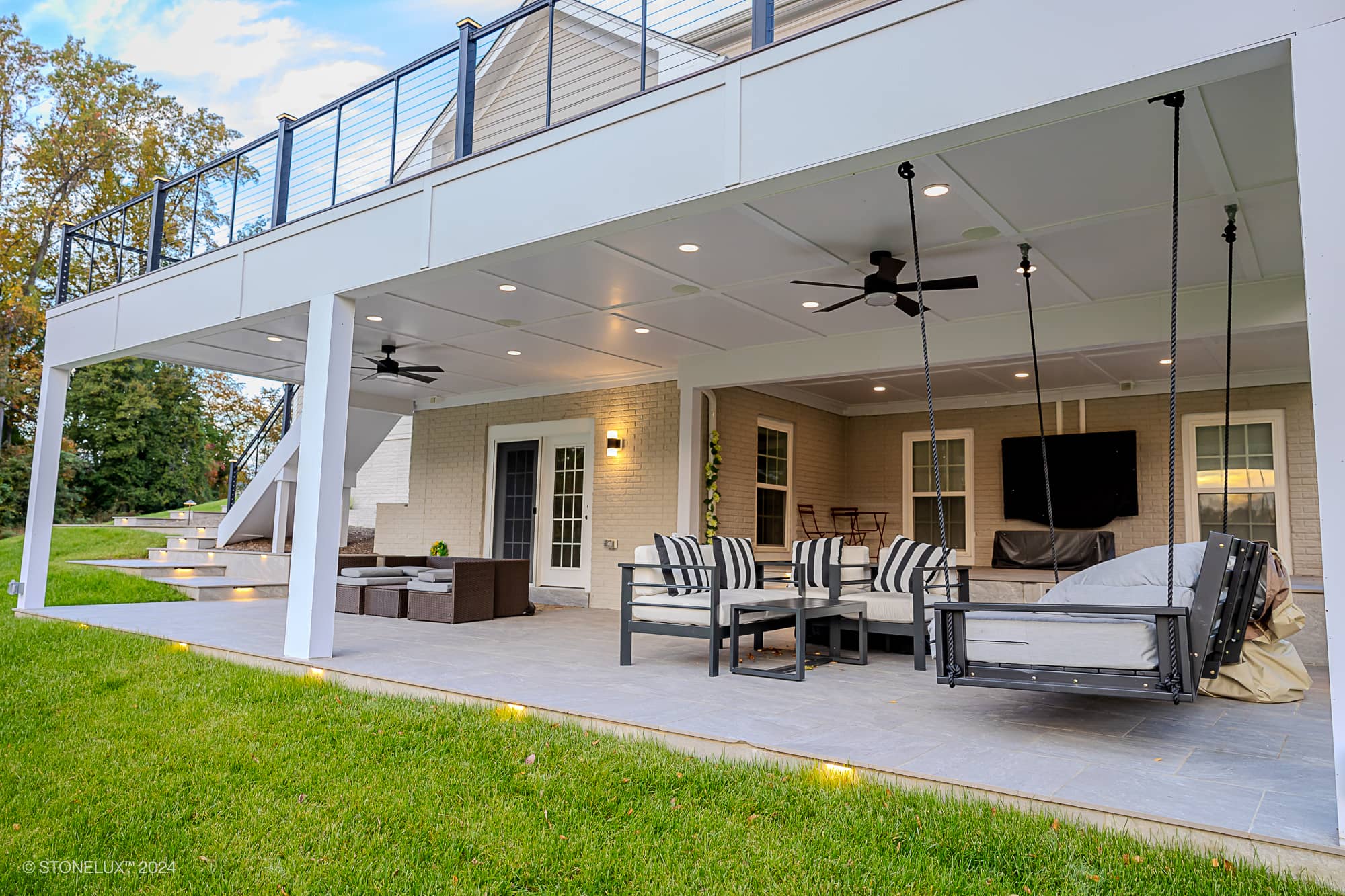 A modern two-story deck attached to a beige house features outdoor furniture, lounge chairs on the upper level, and a seating area below with built-in lighting, all overlooking a Rasa Grey: Porcelain Paver Patio in Fairfax and lush green lawn.