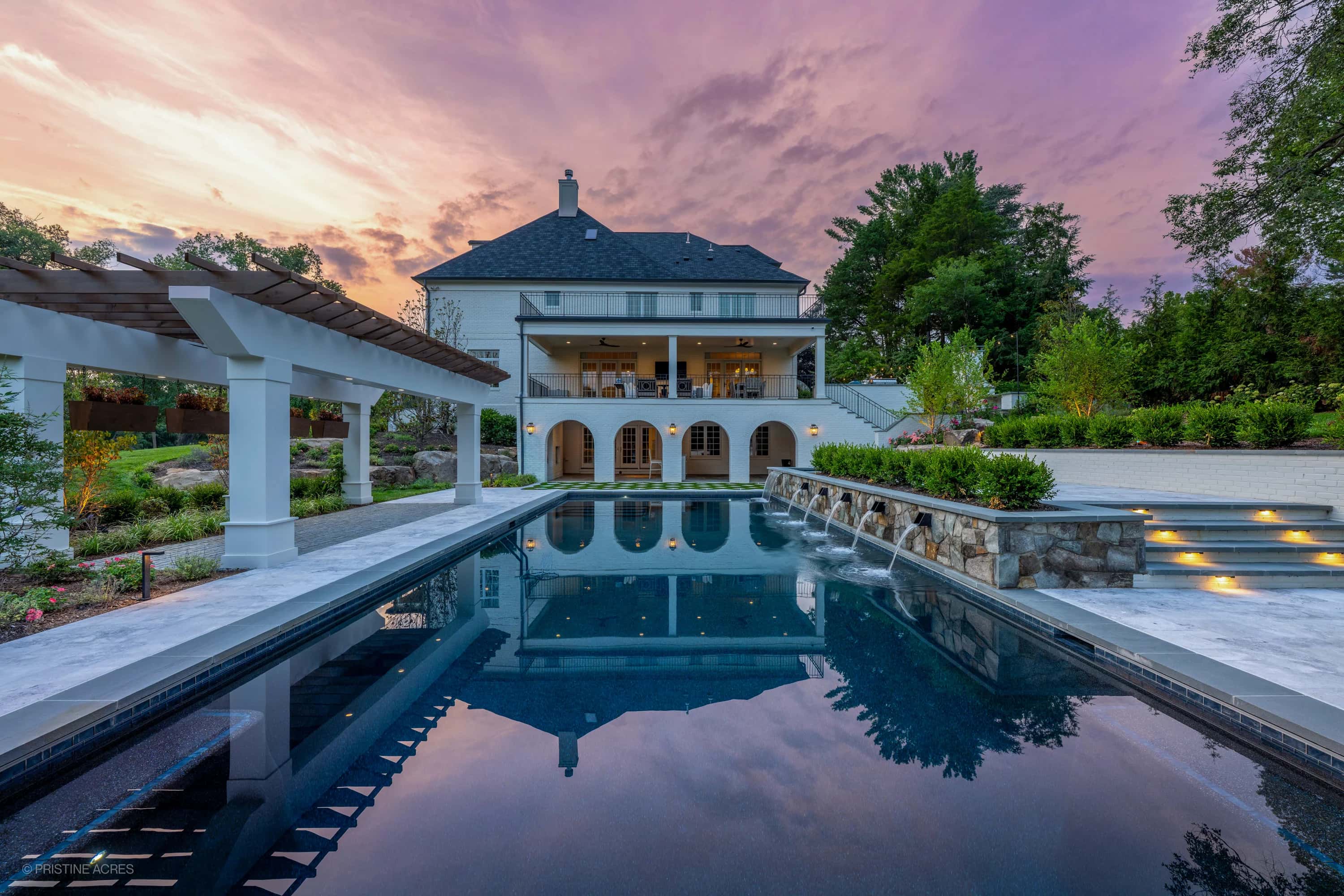 Luxurious Tundra Grey Marble pool deck with Bluestone coping in McLean, VA. Modern design featuring a reflective pool, cascading water features, and lush landscaping. Elegant pergola and ambient lighting create a serene backyard oasis.