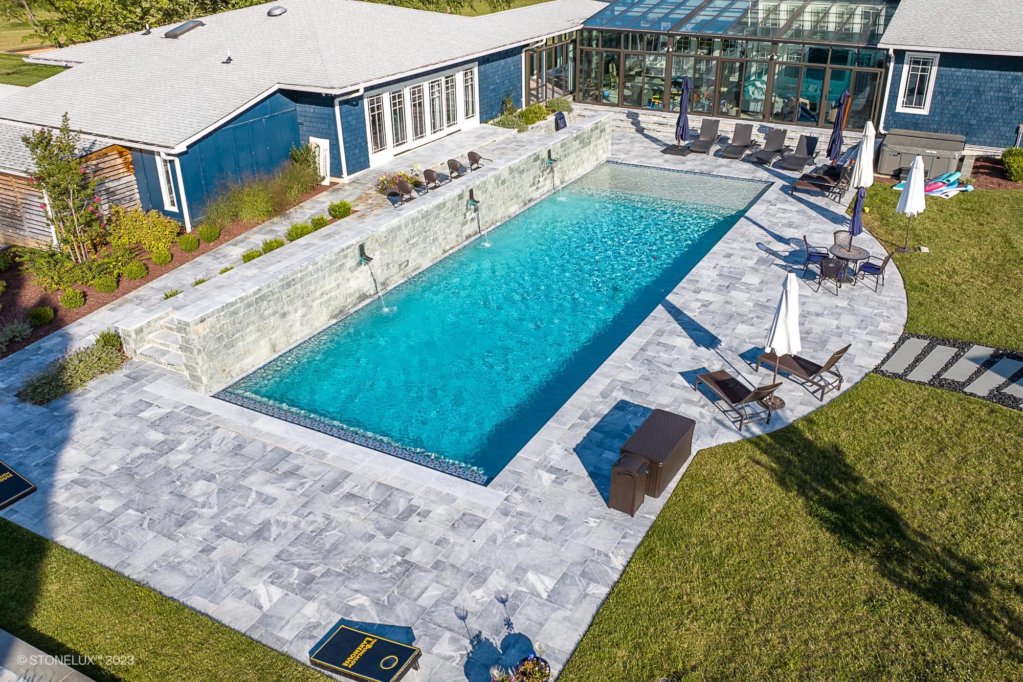 Aerial view of Blue Haven: The Grey and Blue Sandblasted Marble Pool Project, featuring a rectangular swimming pool with three water features, gray stone tiles, lounge chairs, umbrellas, and a modern house with a large glass sunroom.