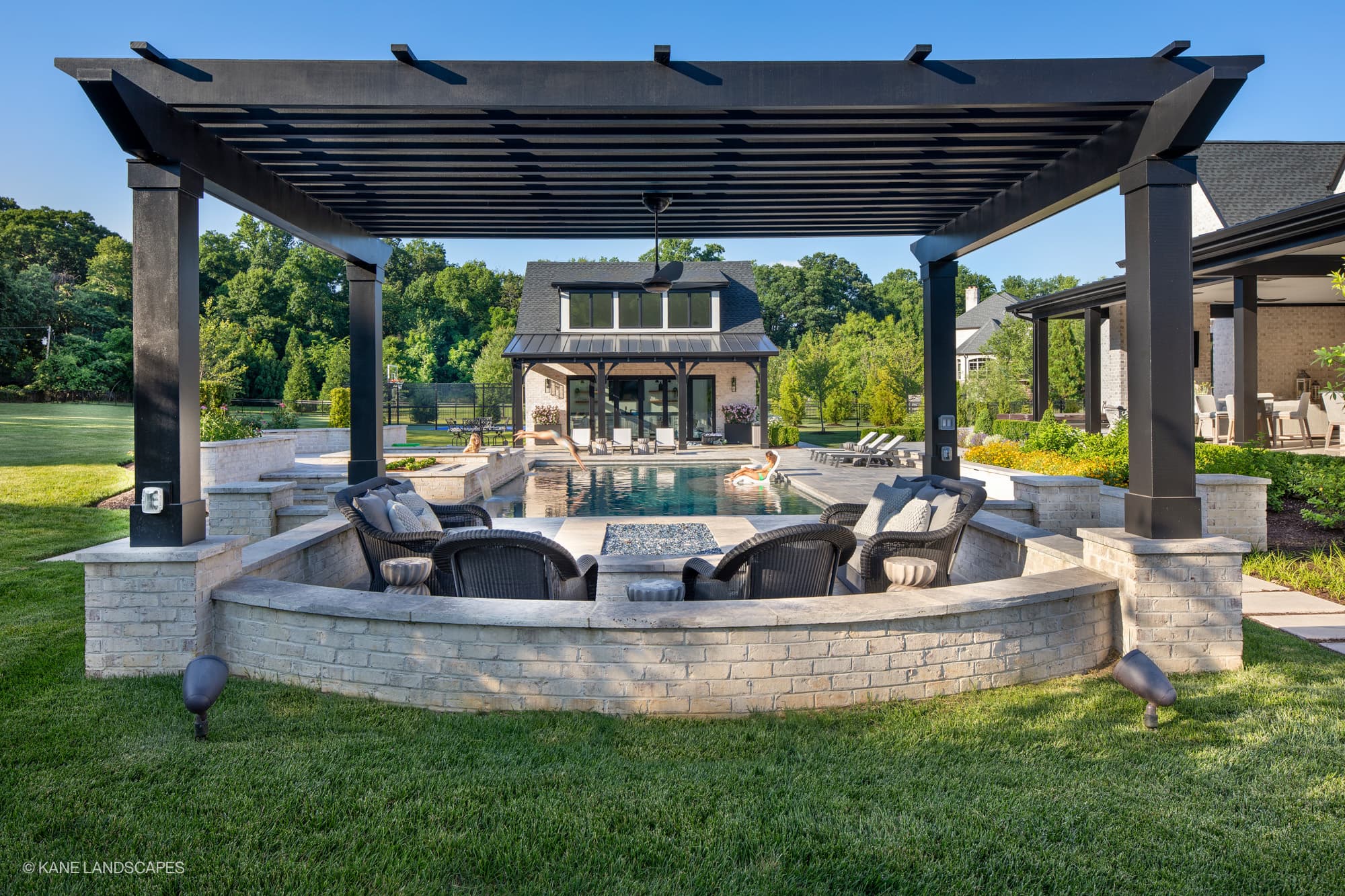 Modern Oasis at Grenata Preserve features Silver Travertine Premium pavers in a contemporary Leesburg, VA backyard. The sleek pool reflects the blue sky, surrounded by lush greenery and stylish black pergola. Cozy wicker seating complements the elegant...