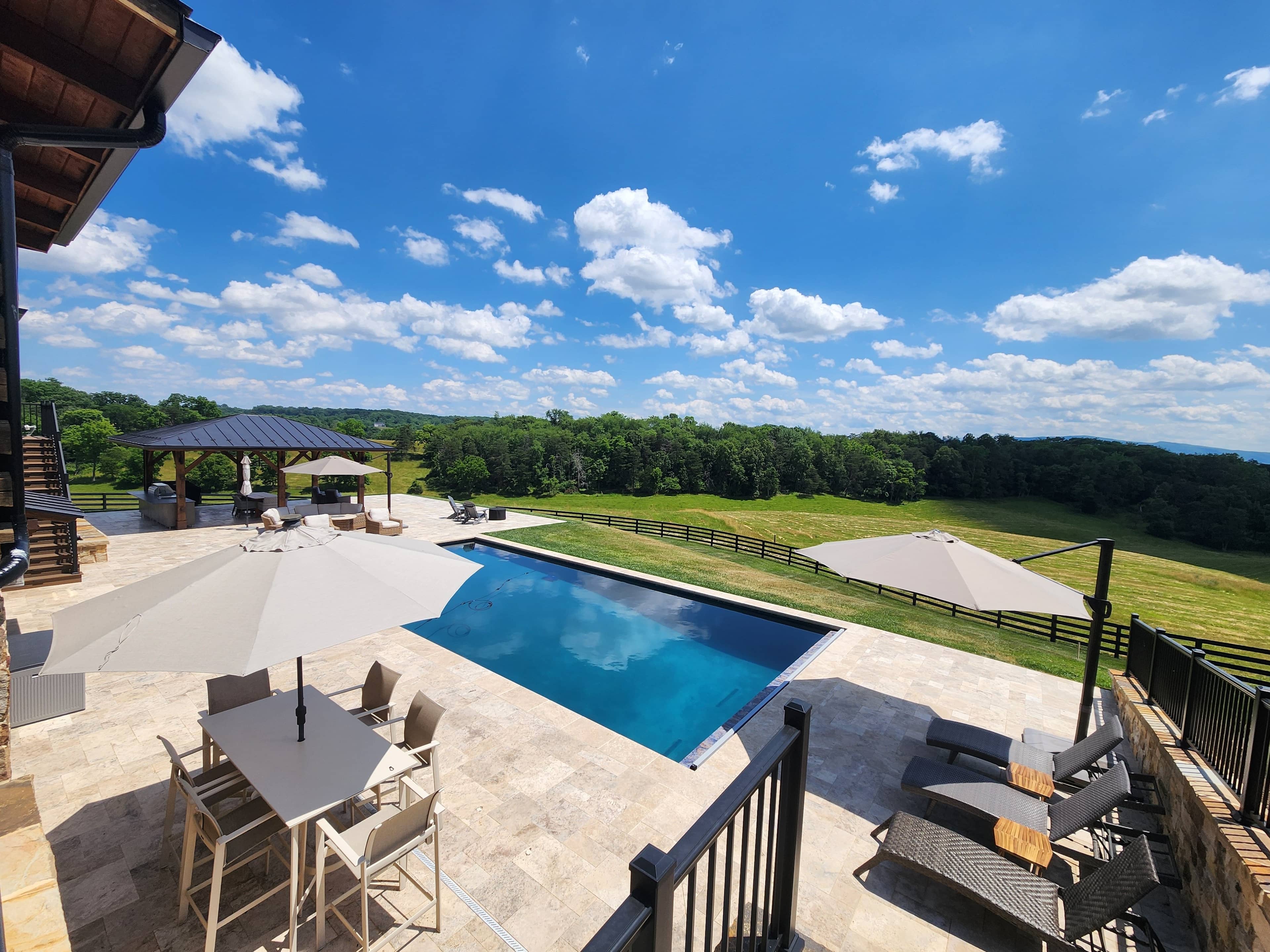 A modern Winchester, VA Pool & Patio Project showcases Philly Travertine Elegance with a rectangular swimming pool, lounge chairs, umbrellas, dining table, and gazebo overlooking a grassy field and trees beneath a blue, cloud-dotted sky.