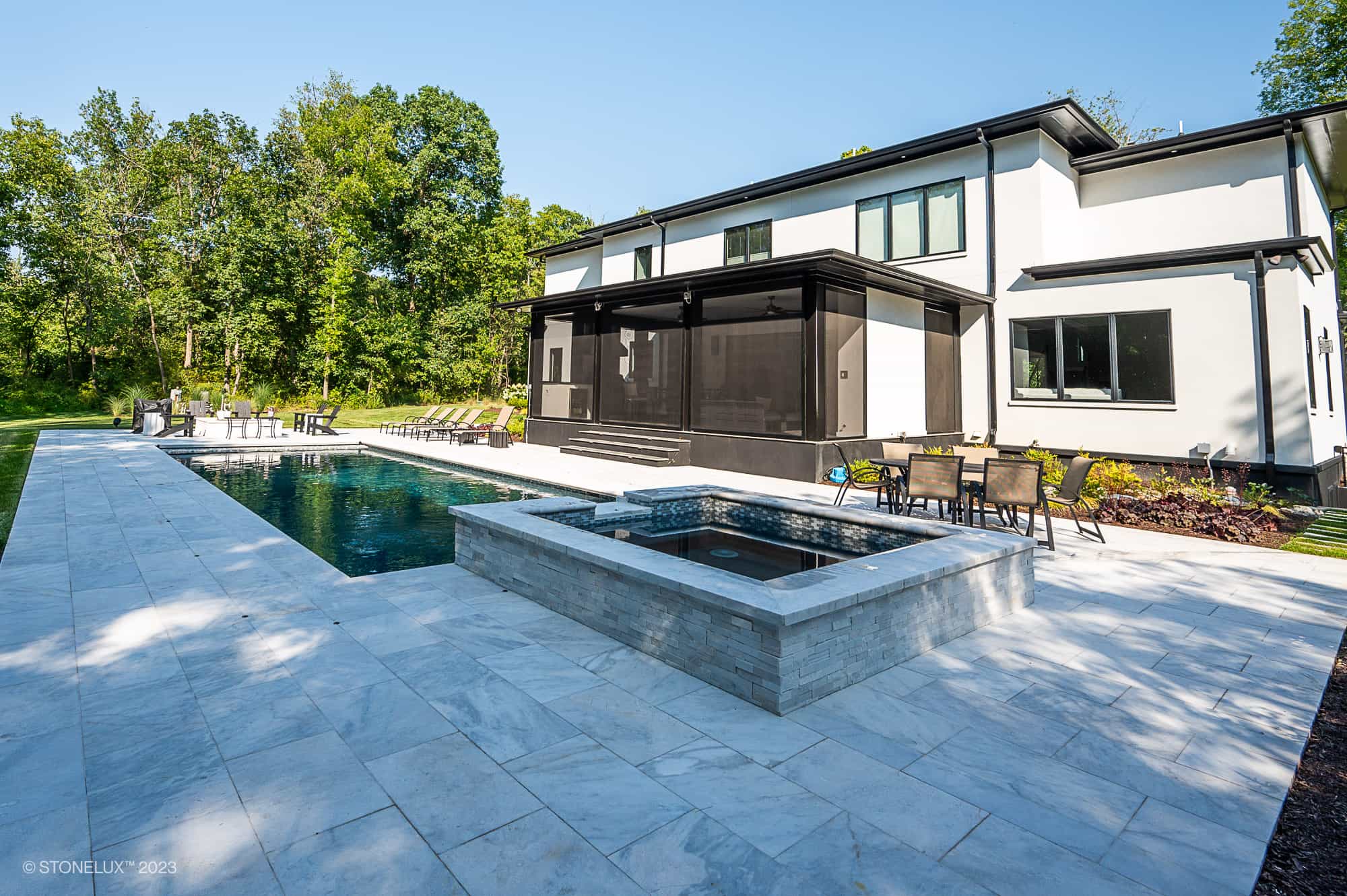 Modern backyard featuring a rectangular swimming pool and hot tub, with lounge chairs and patio tables. The Afyon Elegance: Modern White Marvel with Black Accents Poolside complements the two-story house surrounded by lush trees and greenery.