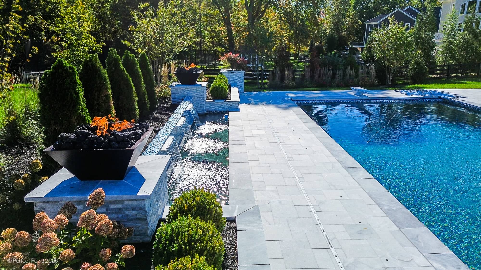 Aldie Serenity – Afyon White & Arctic Grey Marble Luxury Pool Project: A modern backyard features a rectangular swimming pool with clear water, bordered by tall shrubs, flowerbeds, and decorative fire bowls.