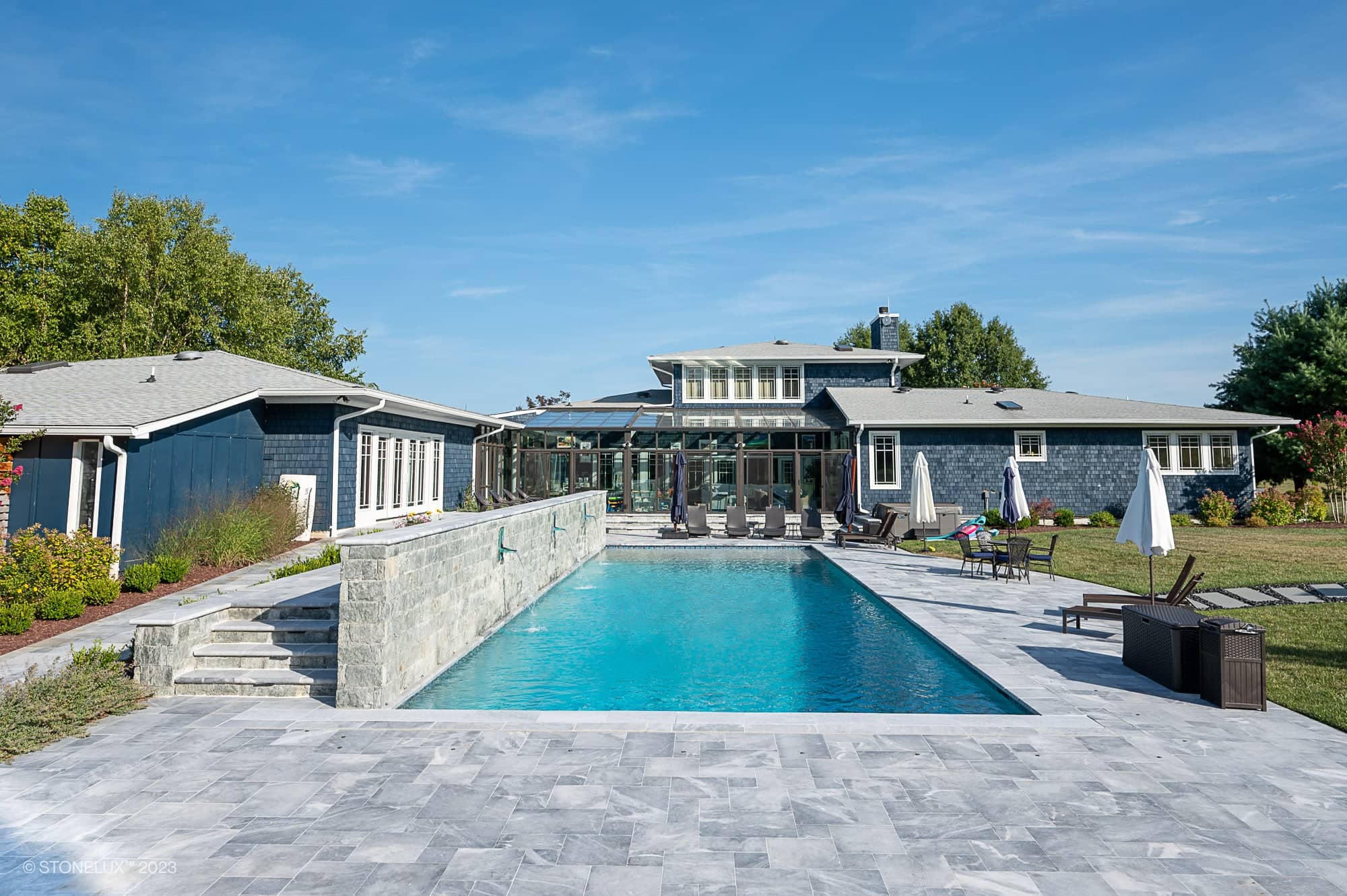 A modern house with large windows and gray siding features Blue Haven: The Grey and Blue Sandblasted Marble Pool Project—a rectangular outdoor pool surrounded by stone tiles, lounge chairs, umbrellas, and a landscaped yard under a blue sky.